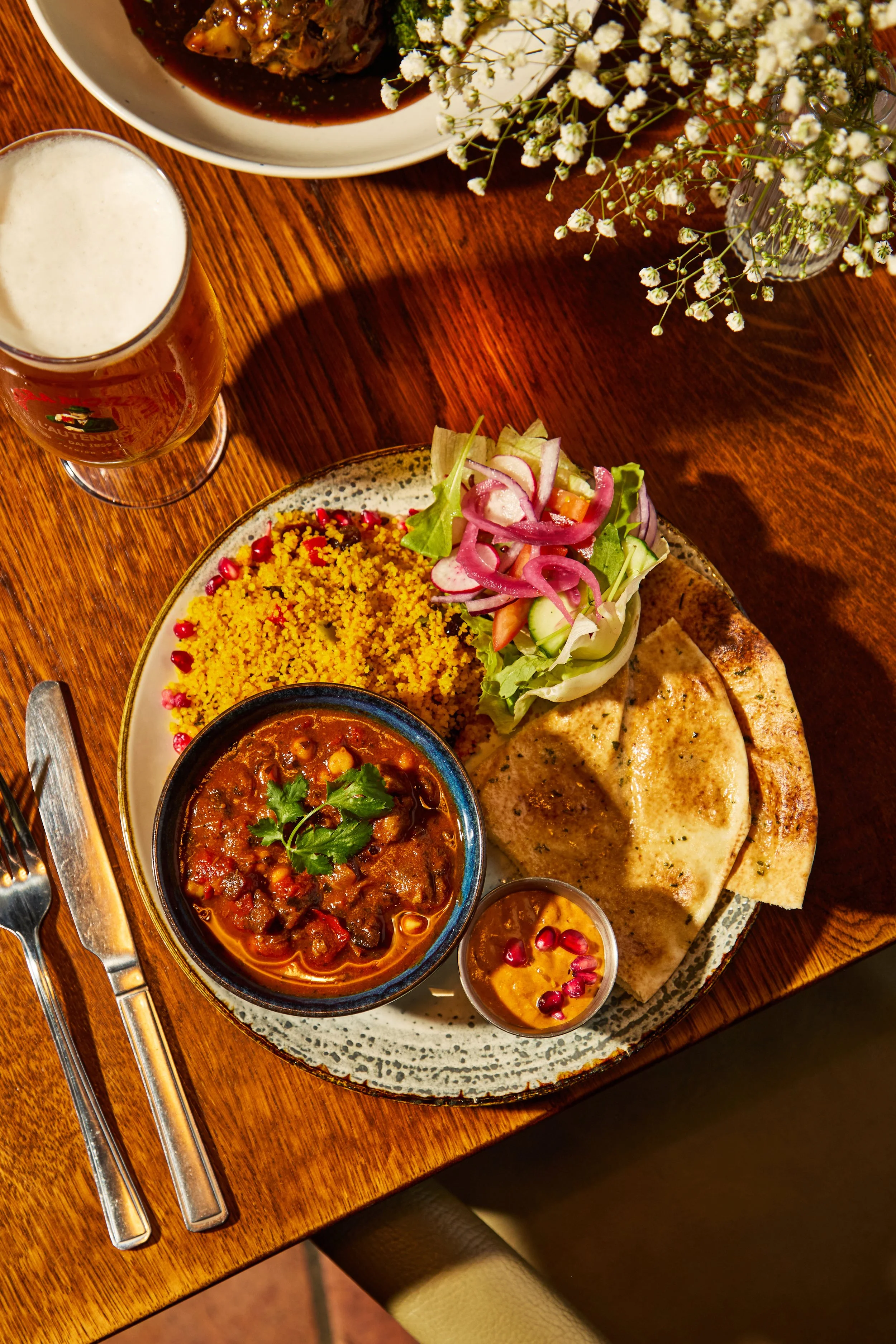 A wooden table with a plate of Mexican food, including a bowl of chili, two folded quesadillas, a side of rice, and a small cup of sauce garnished with pomegranate seeds. There is also a glass of beer and a small bowl of soup, along with a vase of wh