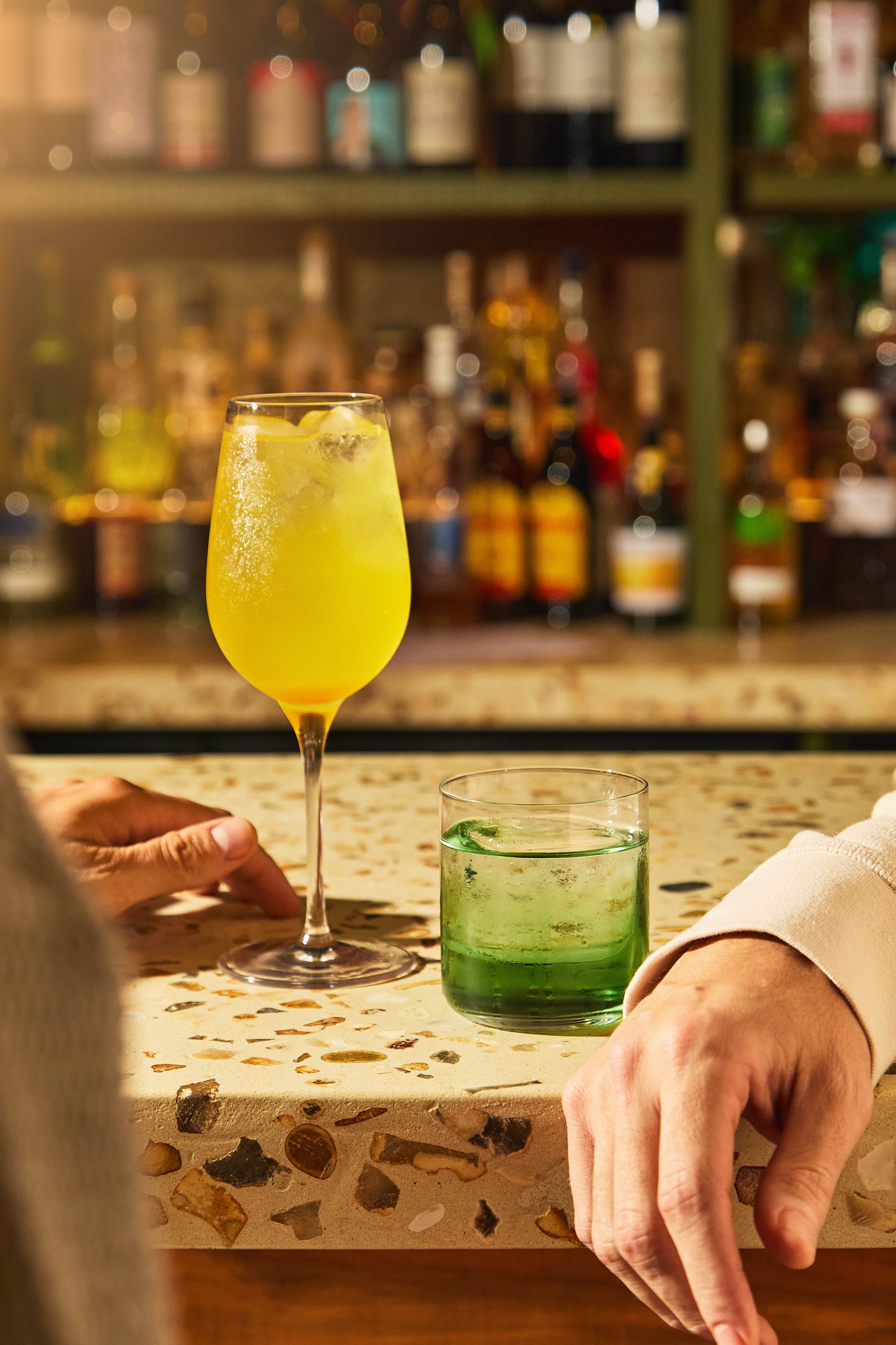 A yellow cocktail in a tall glass with ice and lemon slices, held by a person's hand, on a bar counter with a green drink and blurred liquor bottles in the background.