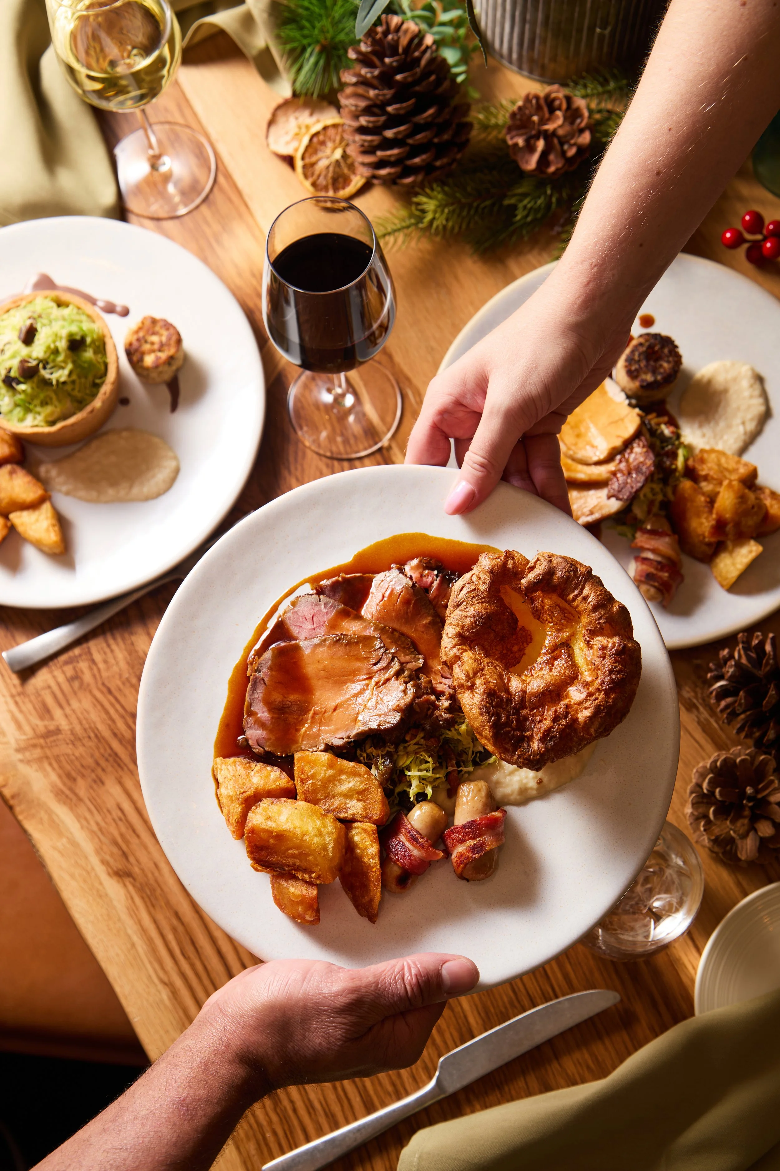 A person holding a plate of Thanksgiving food, including slices of turkey with gravy, stuffing, and potato rolls, over a wooden table set with additional plates of food, wine glasses, and holiday decorations.