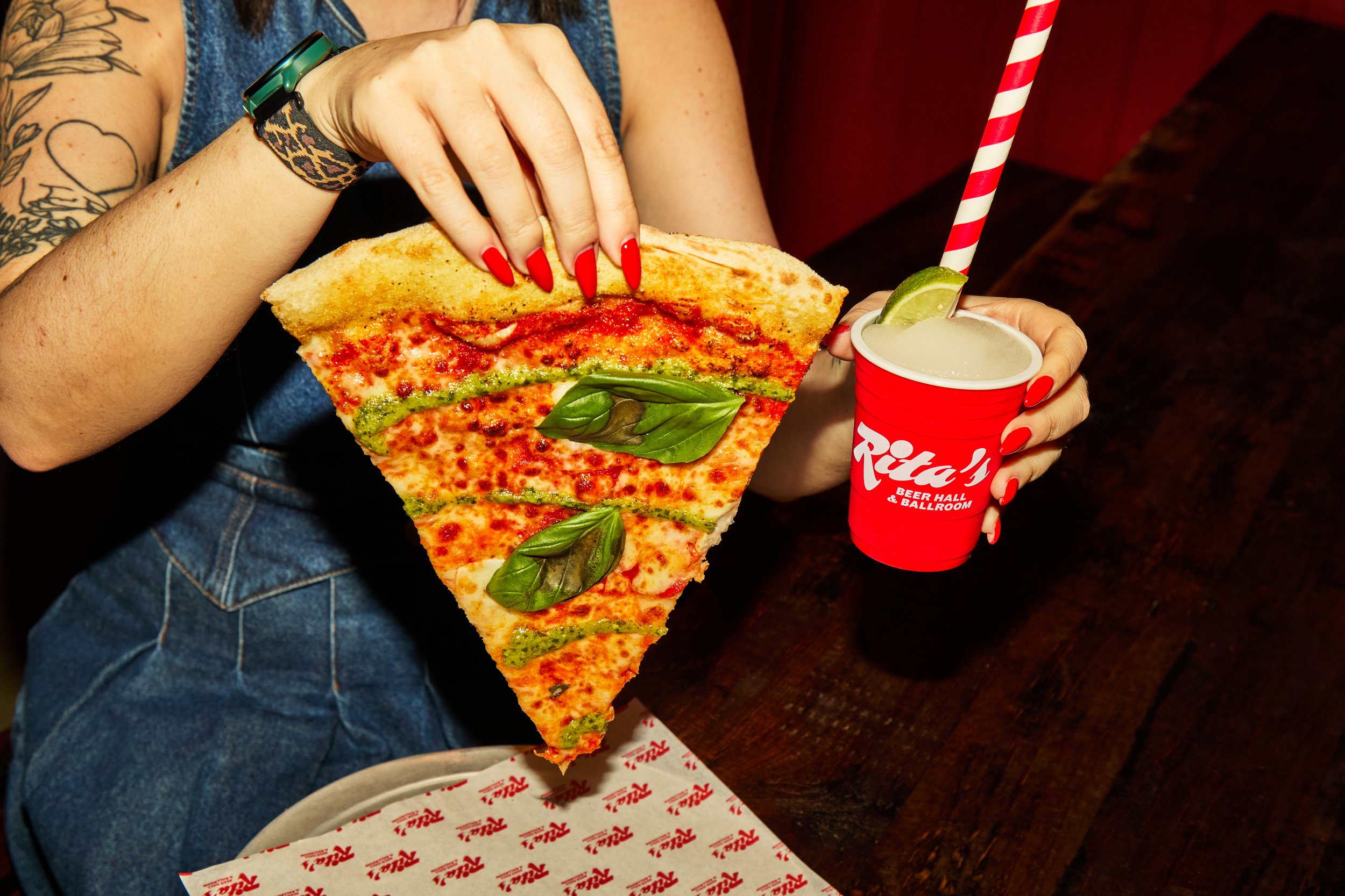 Person holding a large slice of pizza with basil leaves, and a red cup with a straw and lime wedge, seated at a table.