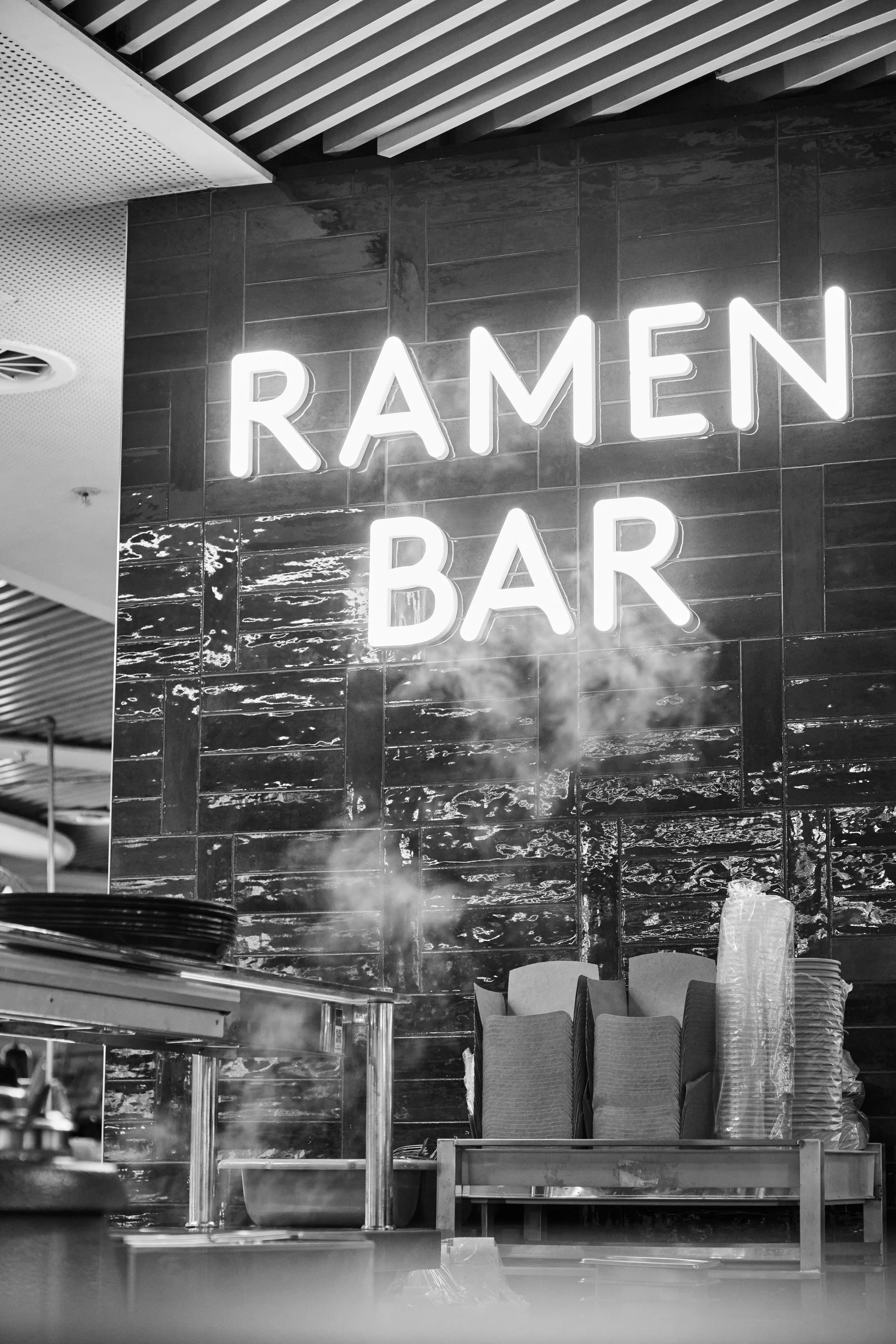 Neon sign reads 'Ramen Bar' on a tiled wall inside a restaurant, with steam rising from kitchen utensils in the foreground.