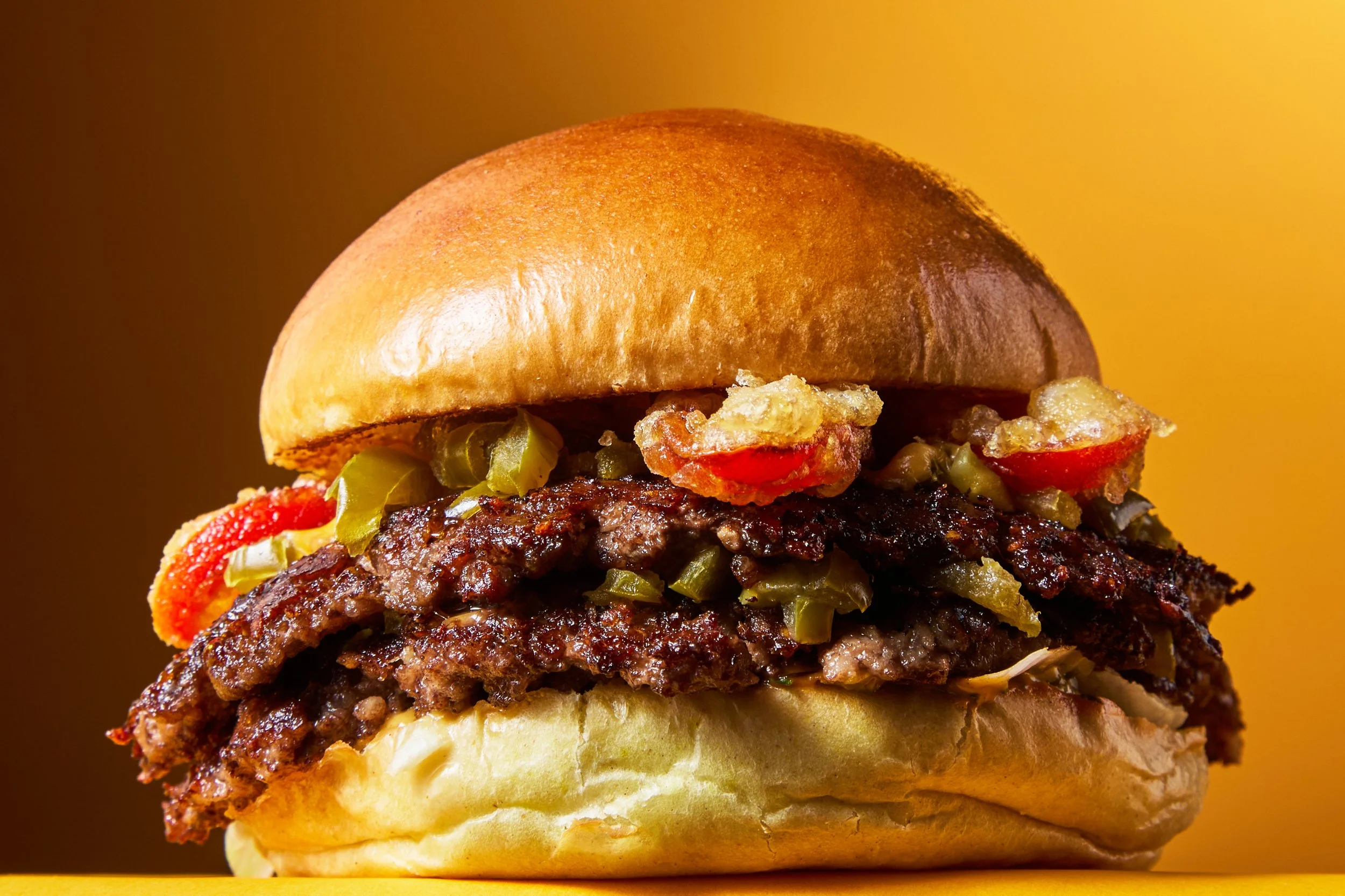 Close-up of a hamburger with a shiny bun, grilled beef patties, pickles, lettuce, and sliced tomatoes.