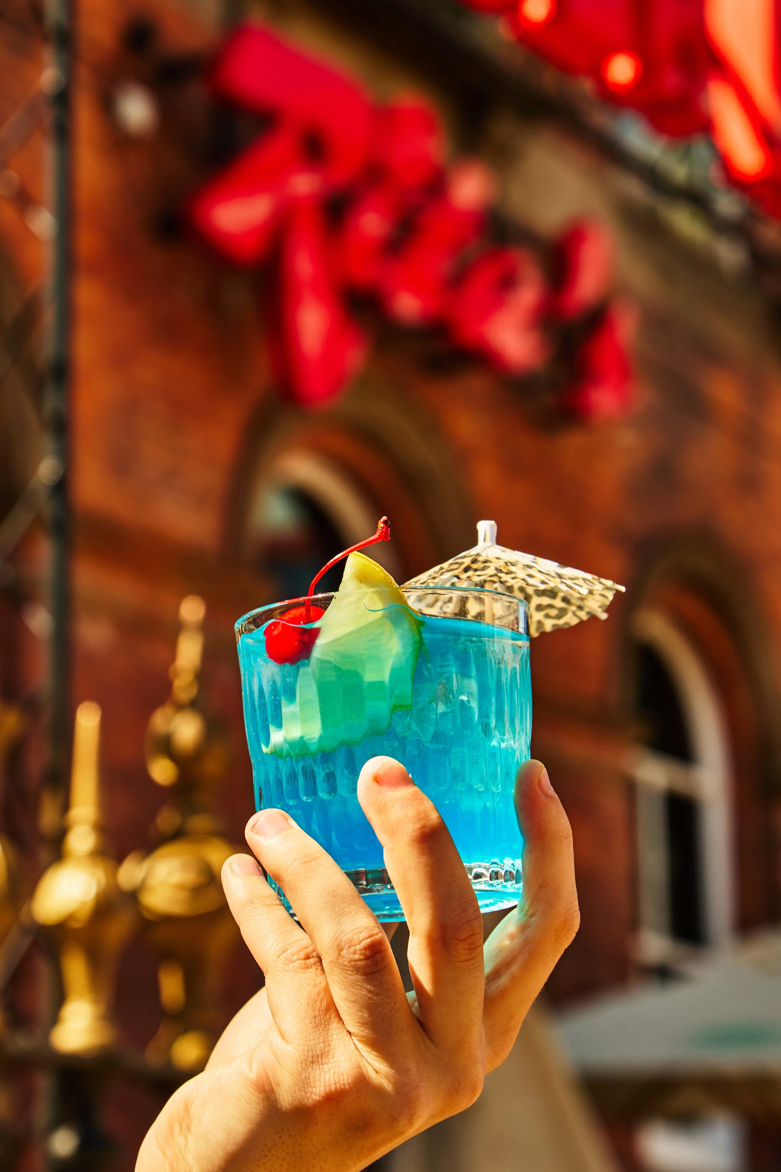 A hand holding a blue tropical cocktail with a cherry, pineapple slice, and umbrella against a blurred background of red decorations.