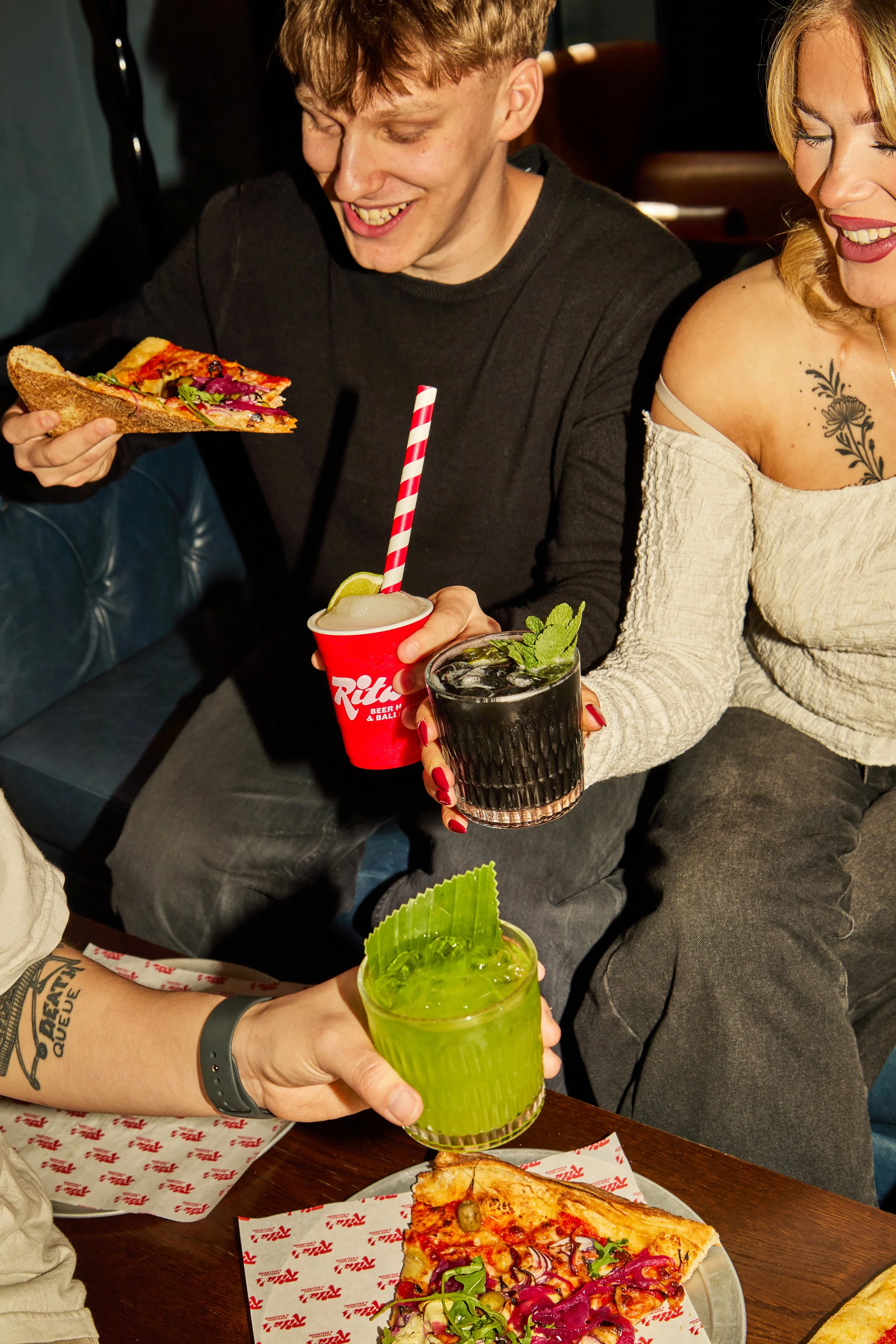 Two people enjoying a meal with drinks and pizza in a restaurant. One person holds a green drink with mint, another has a red drink with a straw, and there is a slice of pizza on a plate.