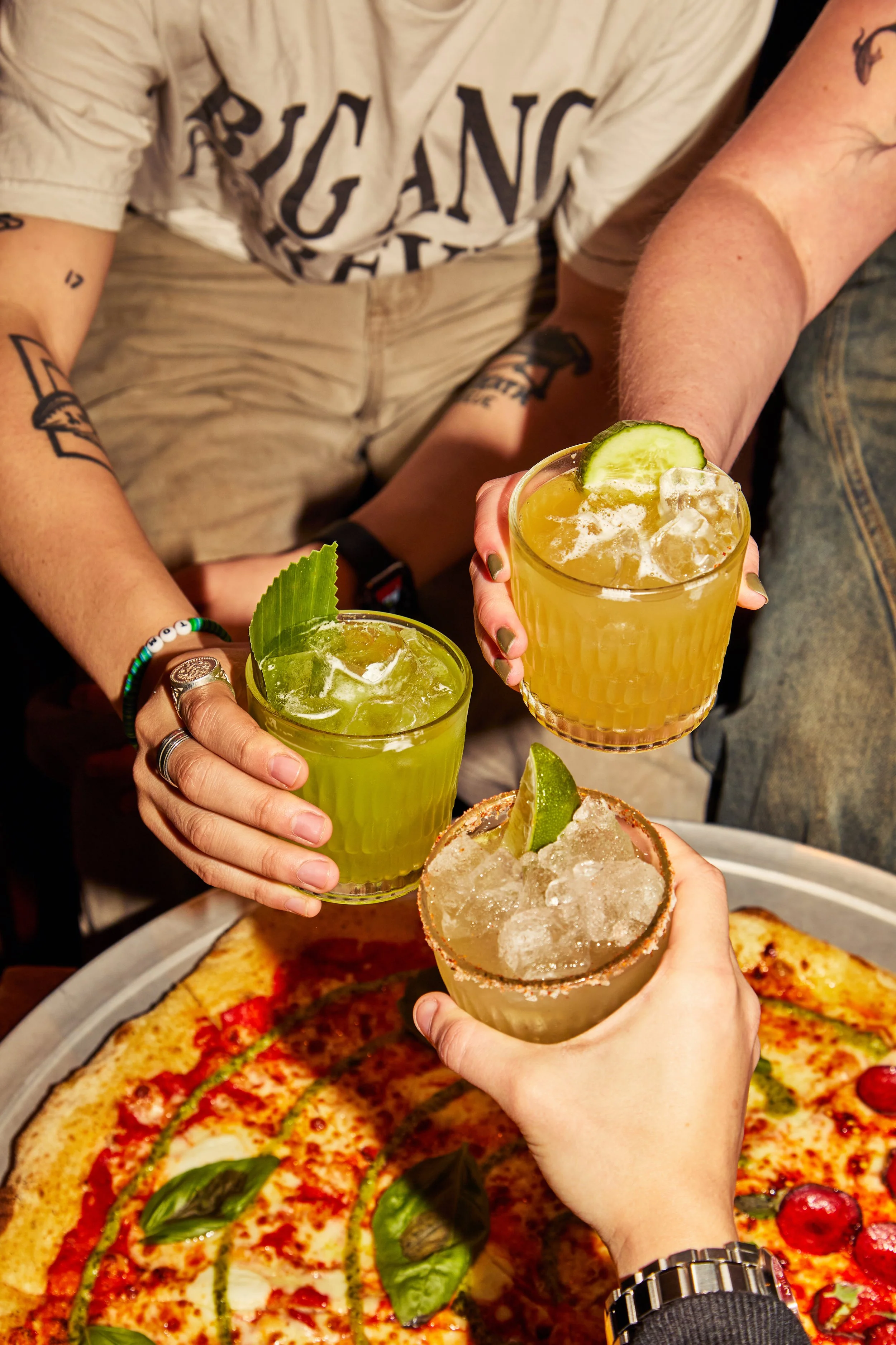 Several hands holding up various cocktails over a large pizza with toppings including basil, cherry tomatoes, and sauce, in a casual dining setting.