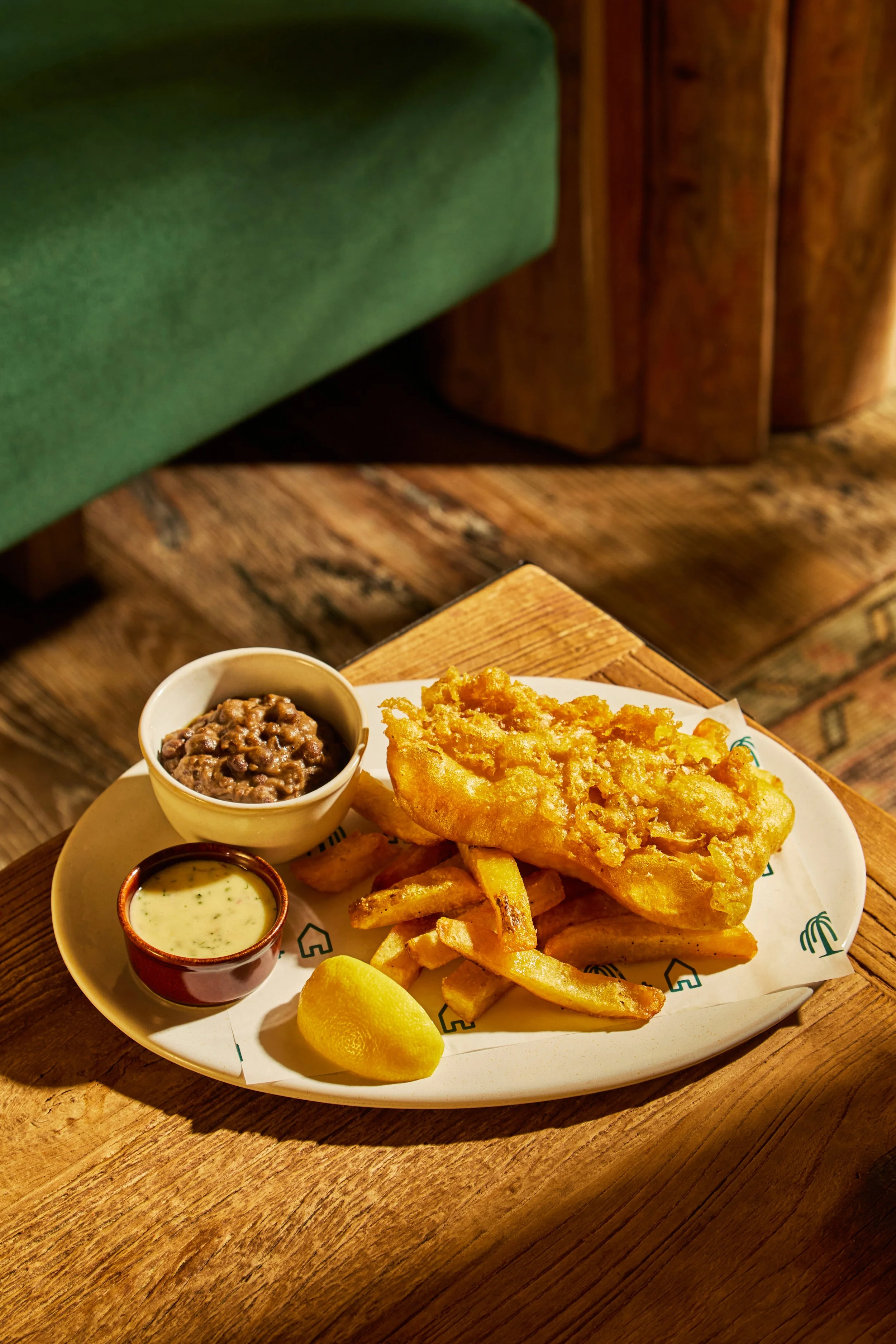 A plate of fish and chips with tartar sauce, baked beans, a lemon wedge, and French fries on a wooden table.