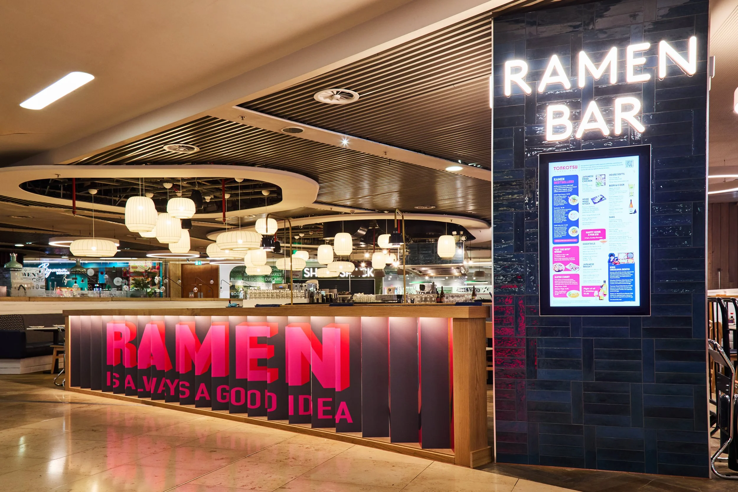 The entrance of a ramen bar with a sign that reads 'RAMEN IS ALWAYS A GOOD IDEA' in pink neon lights. To the right, there is a digital menu display mounted on a dark tiled wall. Inside, the restaurant features hanging paper lantern lights and a moder