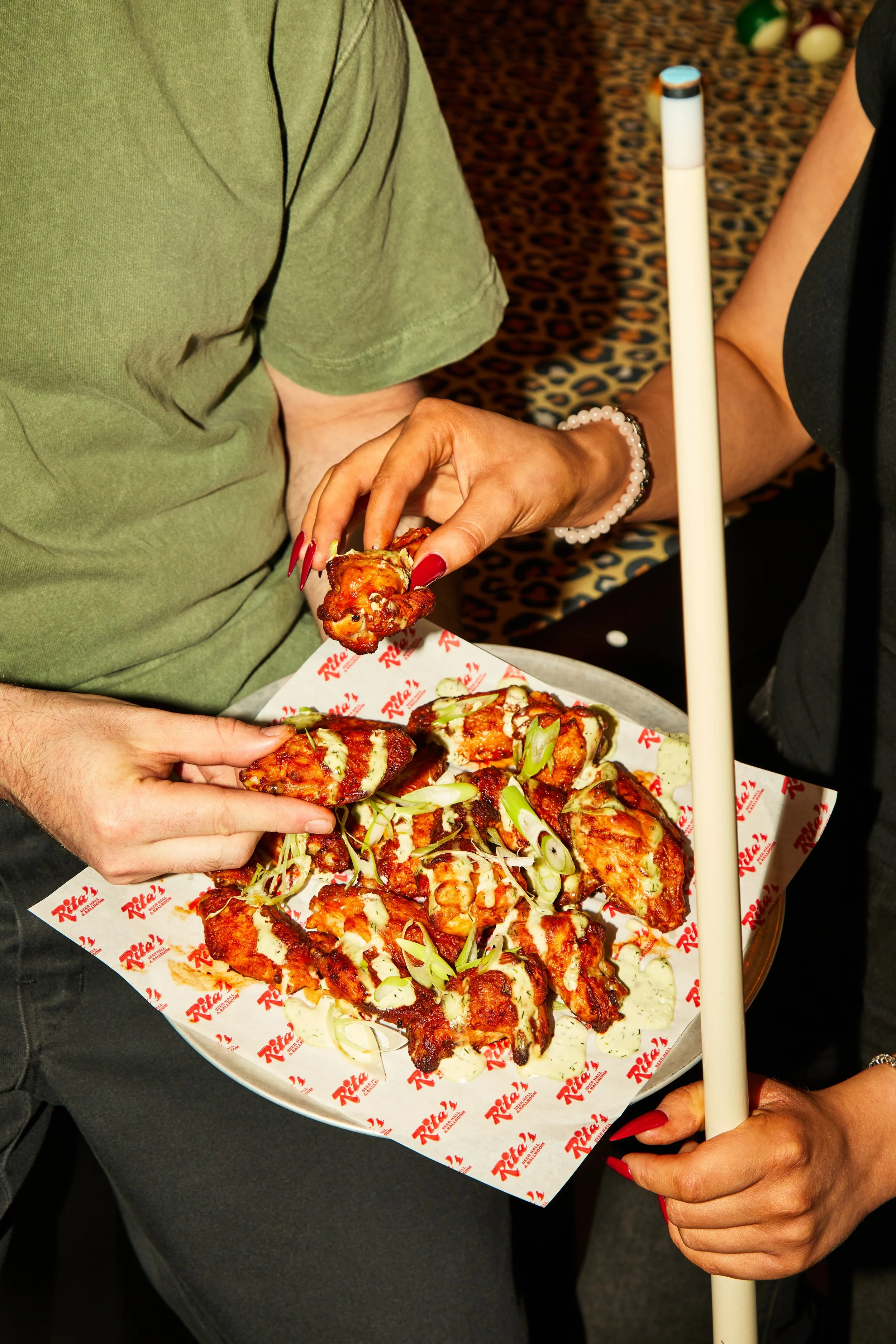 Close-up of two people holding a tray of pepperoni pizza topped with green onions, with one person's hand reaching to grab a slice. The tray has red and white Rita's pizza branding, and the background shows a leopard print carpet and some bowling bal