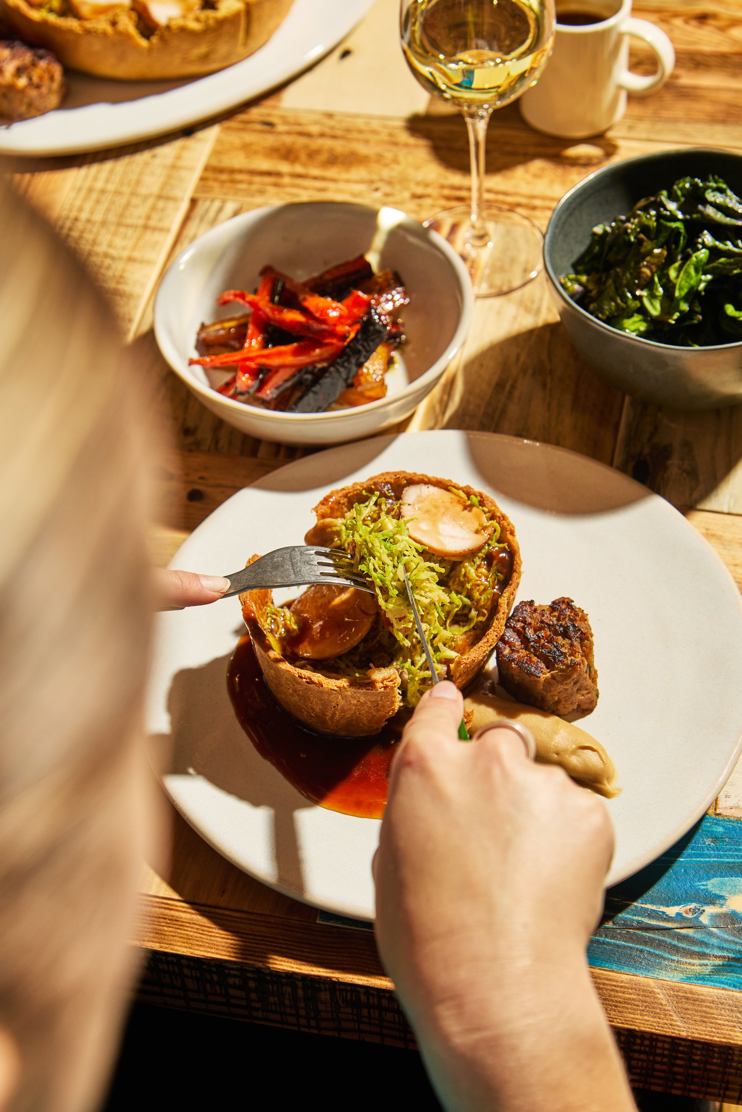 Person holding a fork and knife eating a hollowed-out bread bowl filled with greens and meat, with roasted vegetables and sauces on the plate. A bowl of sautéed greens, a bowl of roasted vegetables, a glass of white wine, and a mug are on a wooden ta