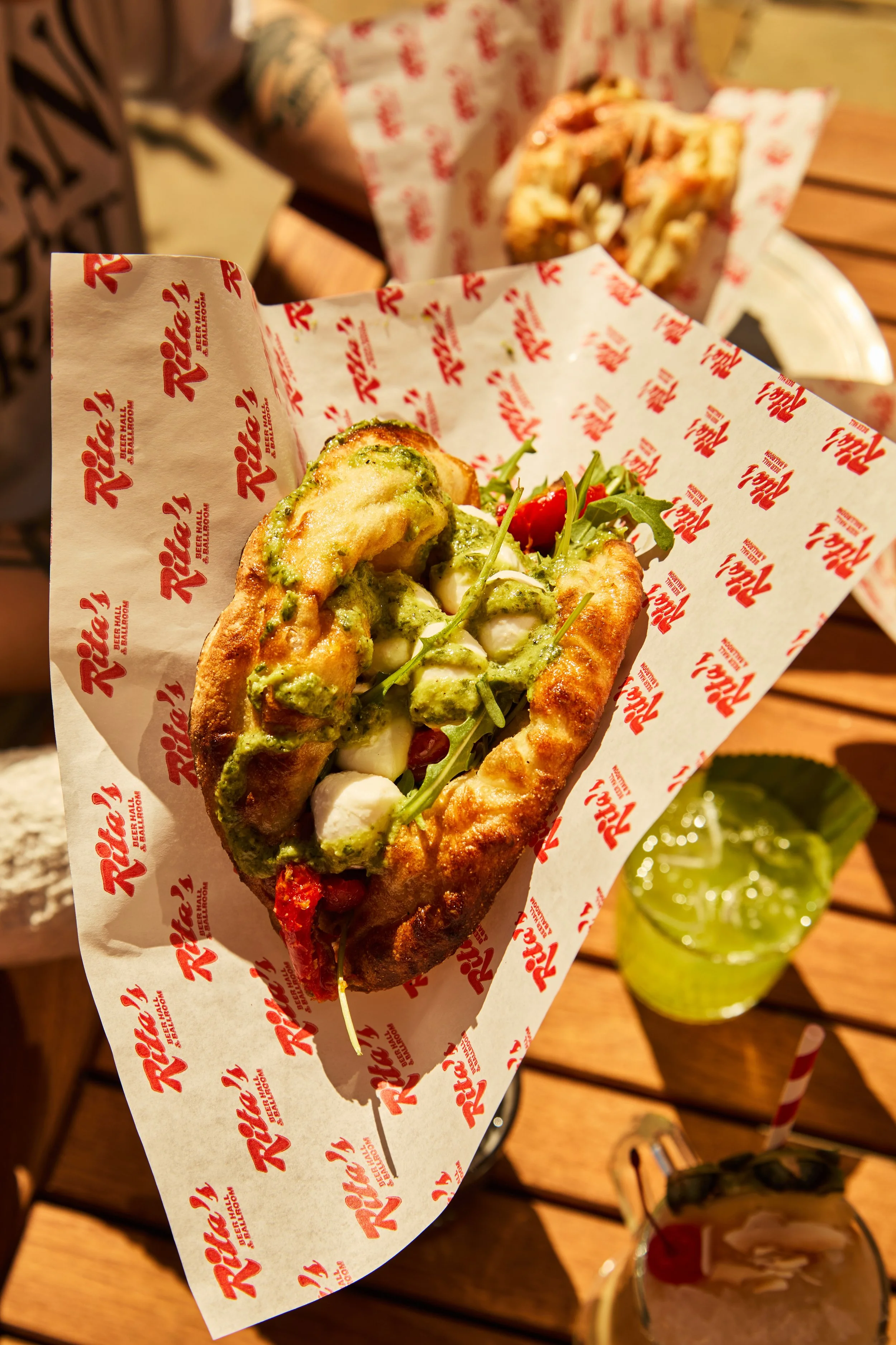 Hot dog topped with green sauce, arugula, cherry tomatoes, and mozzarella cheese, served on branded paper with a lime in the background.