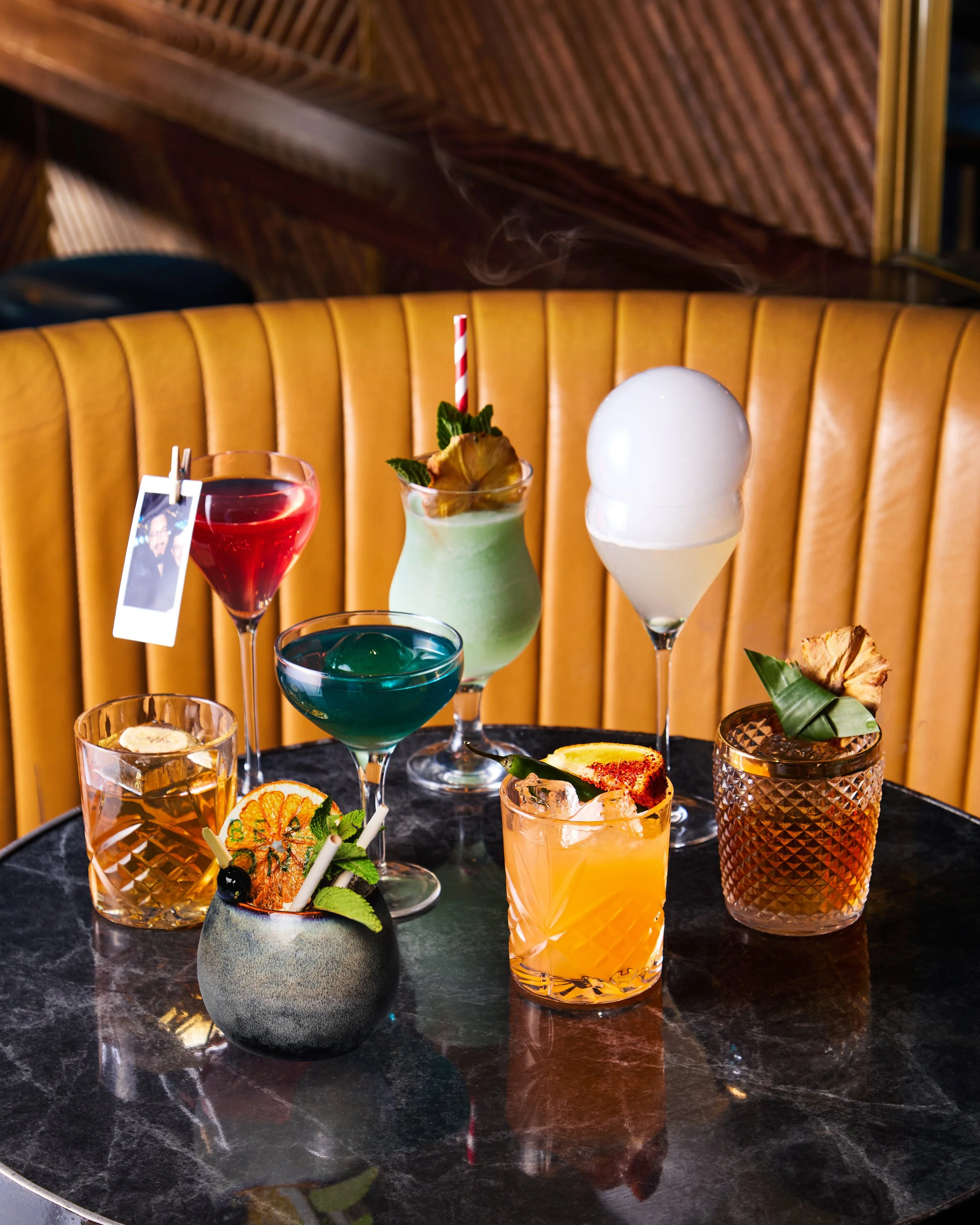 A collection of colorful cocktails on a black marble table with a yellow banquette background.