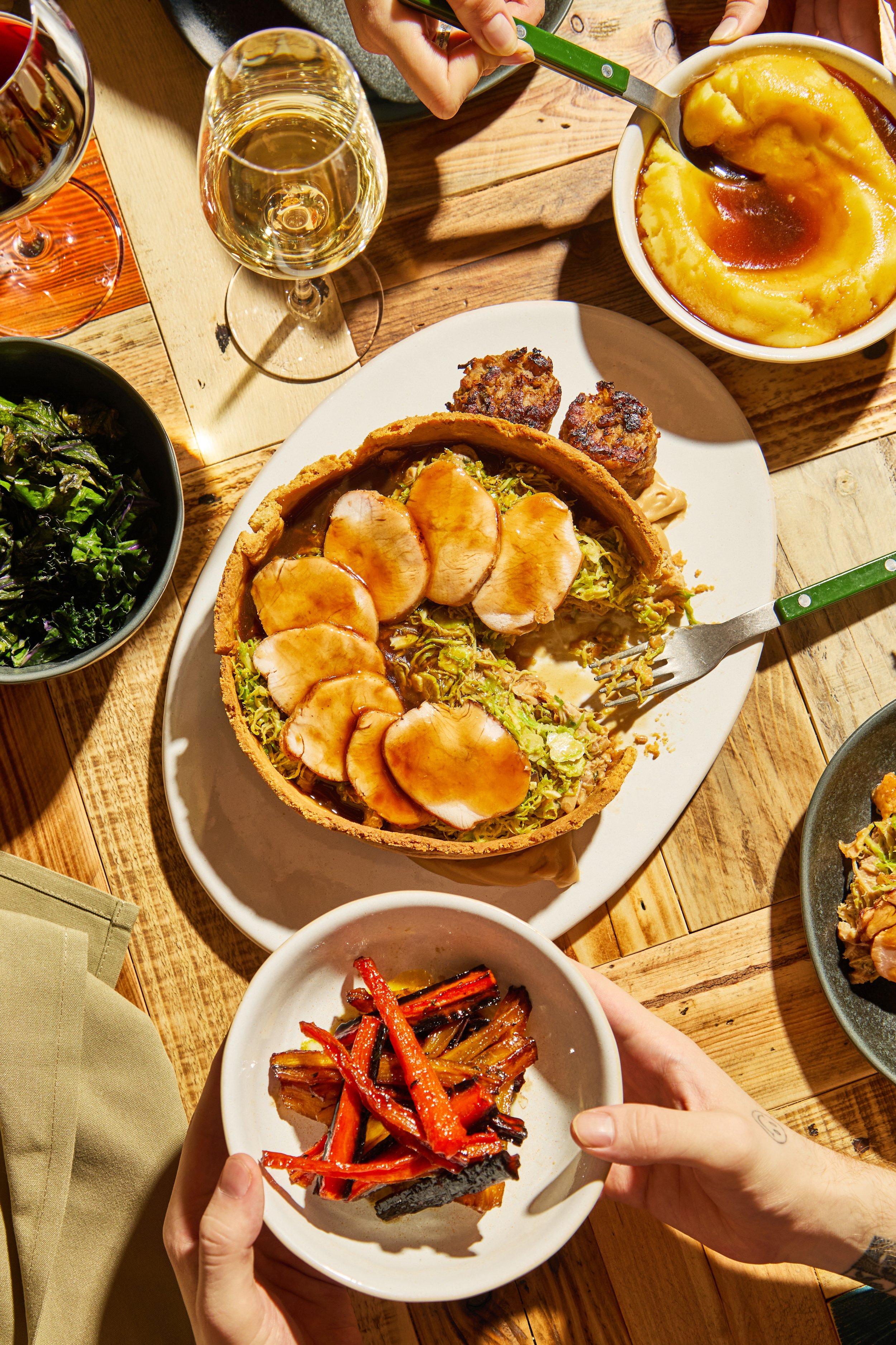 A table with a slice of pumpkin pie topped with whipped cream and caramel, a bowl of mashed potatoes with gravy, a glass of white wine, a bowl of roasted vegetables, a salad, and a plate with roast meat and meatloaf.