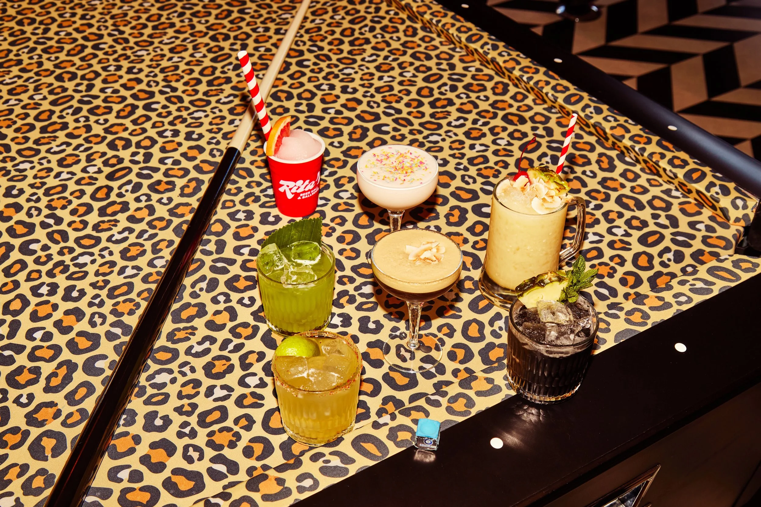 Various colorful cocktails on a table with leopard print tablecloth, surrounded by straws, garnishes, and some bar accessories.