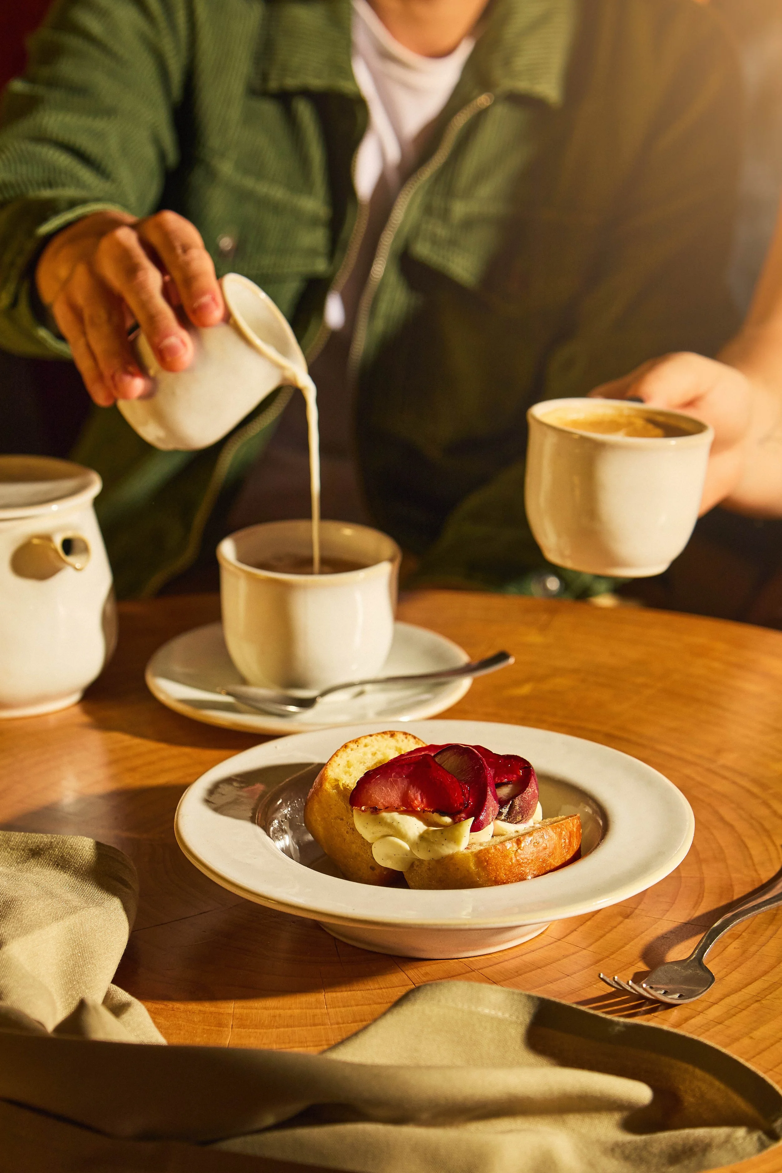 Person pouring cream into a cup of coffee at a table with a dessert and a fork.