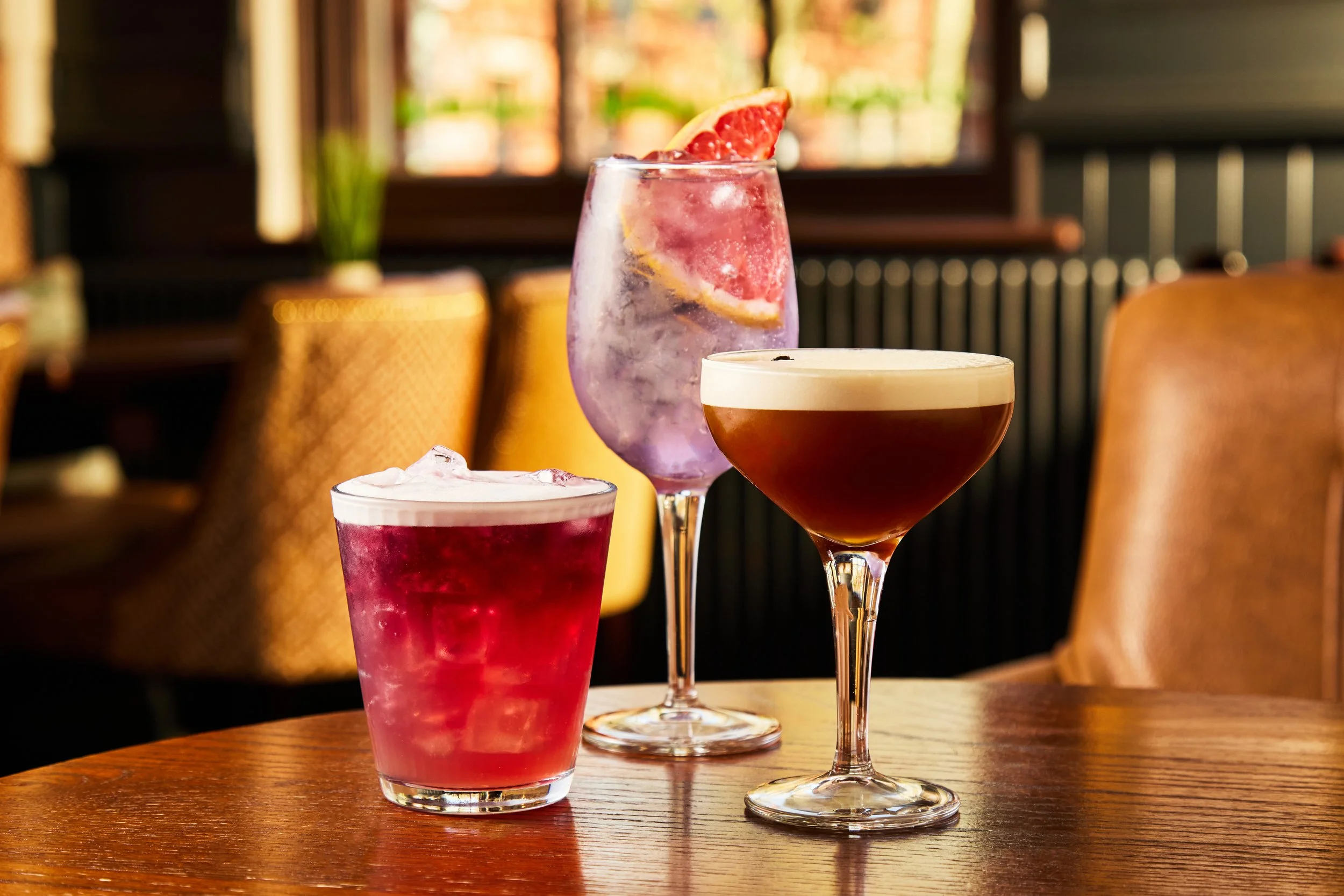 Three colorful cocktails on a wooden table in a cozy restaurant. The center drink is a purple cocktail with a slice of grapefruit garnish, and the two others are red and brown with foam.