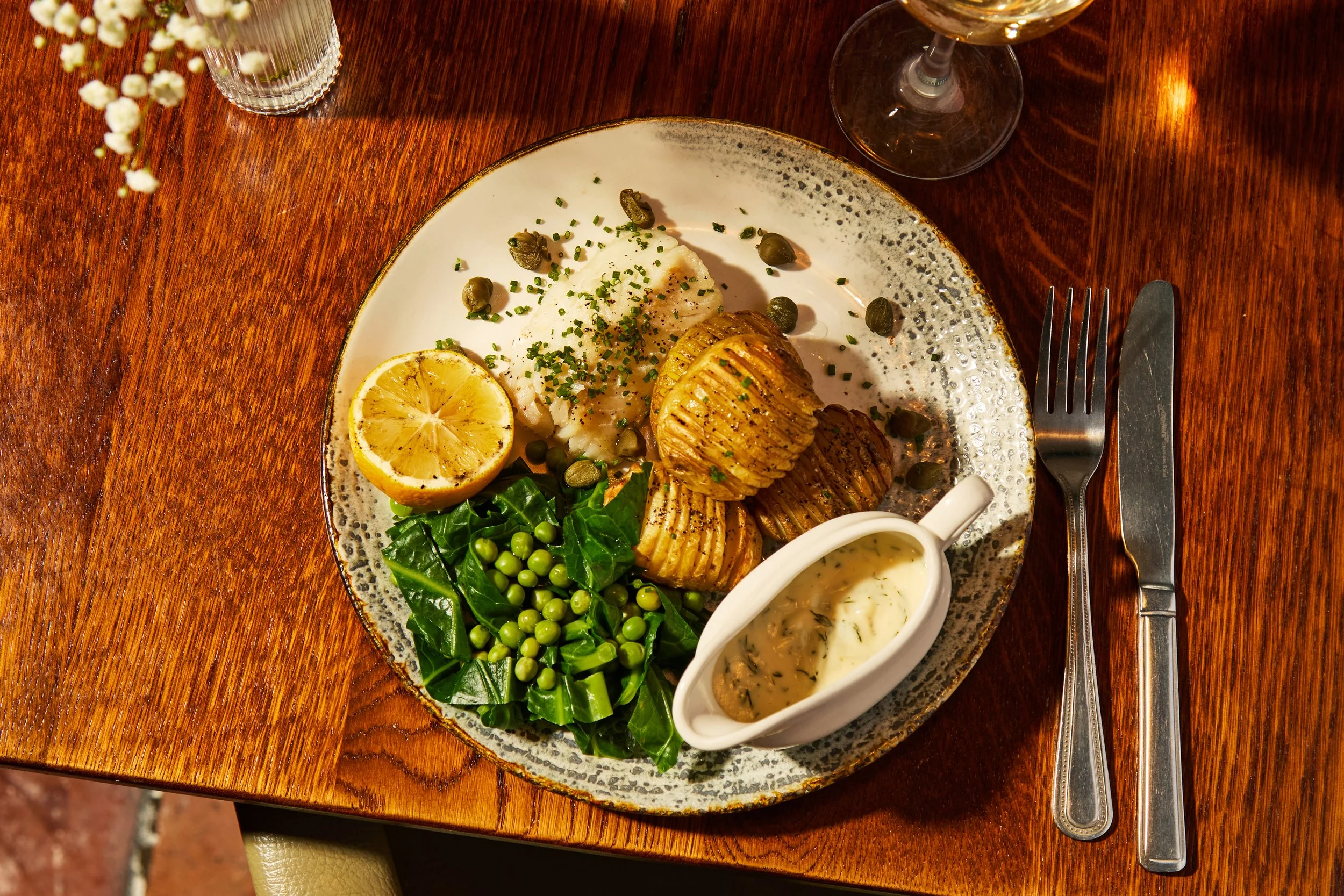 Plate of baked potatoes with chives, a lemon half, mashed potatoes, peas with spinach, and gravy in a gravy boat on a wooden table.
