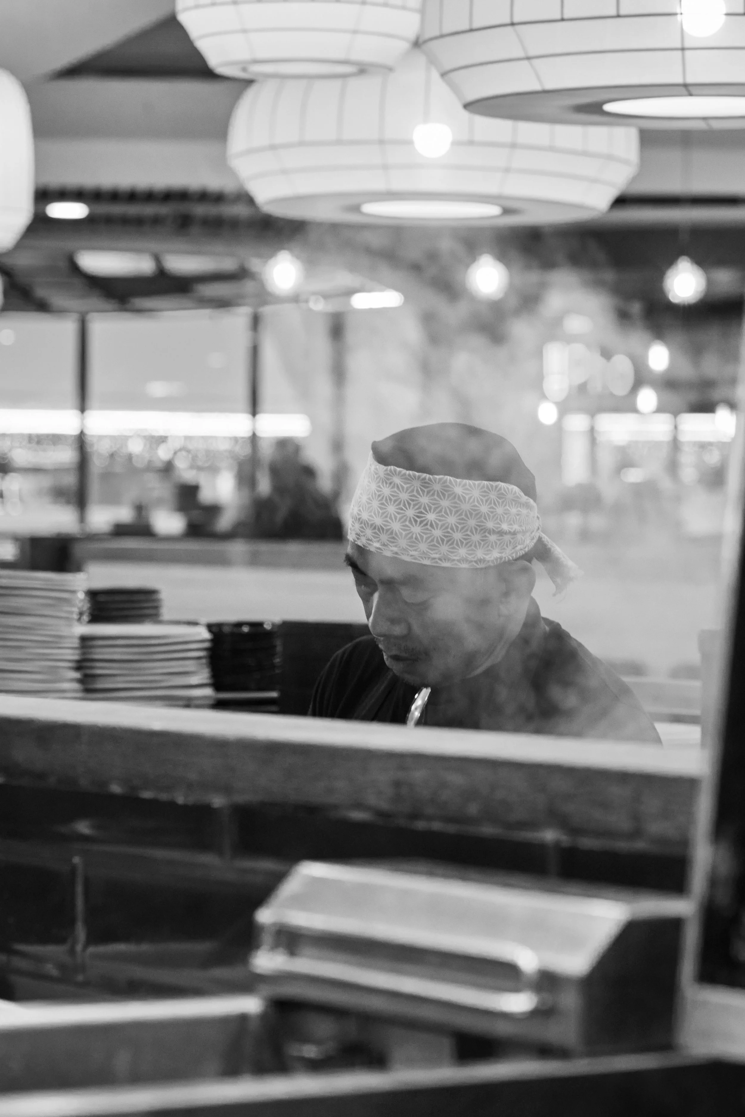 A chef wearing a patterned headband preparing food in a restaurant kitchen, with dishes stacked nearby and illuminated by hanging overhead lights.