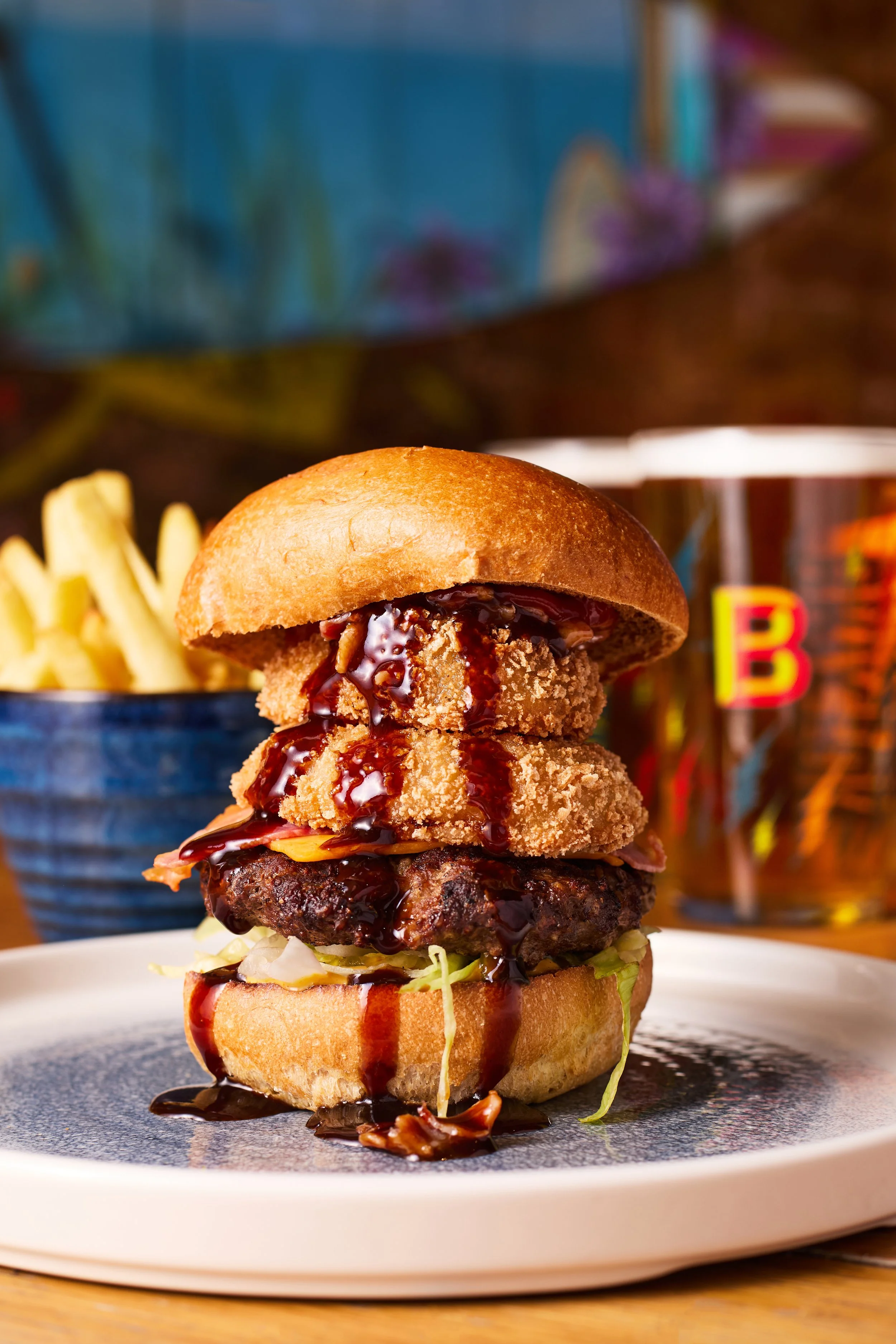 A gourmet burger with crispy fried onion rings and barbecue sauce, served with French fries and a glass of soda in the background.