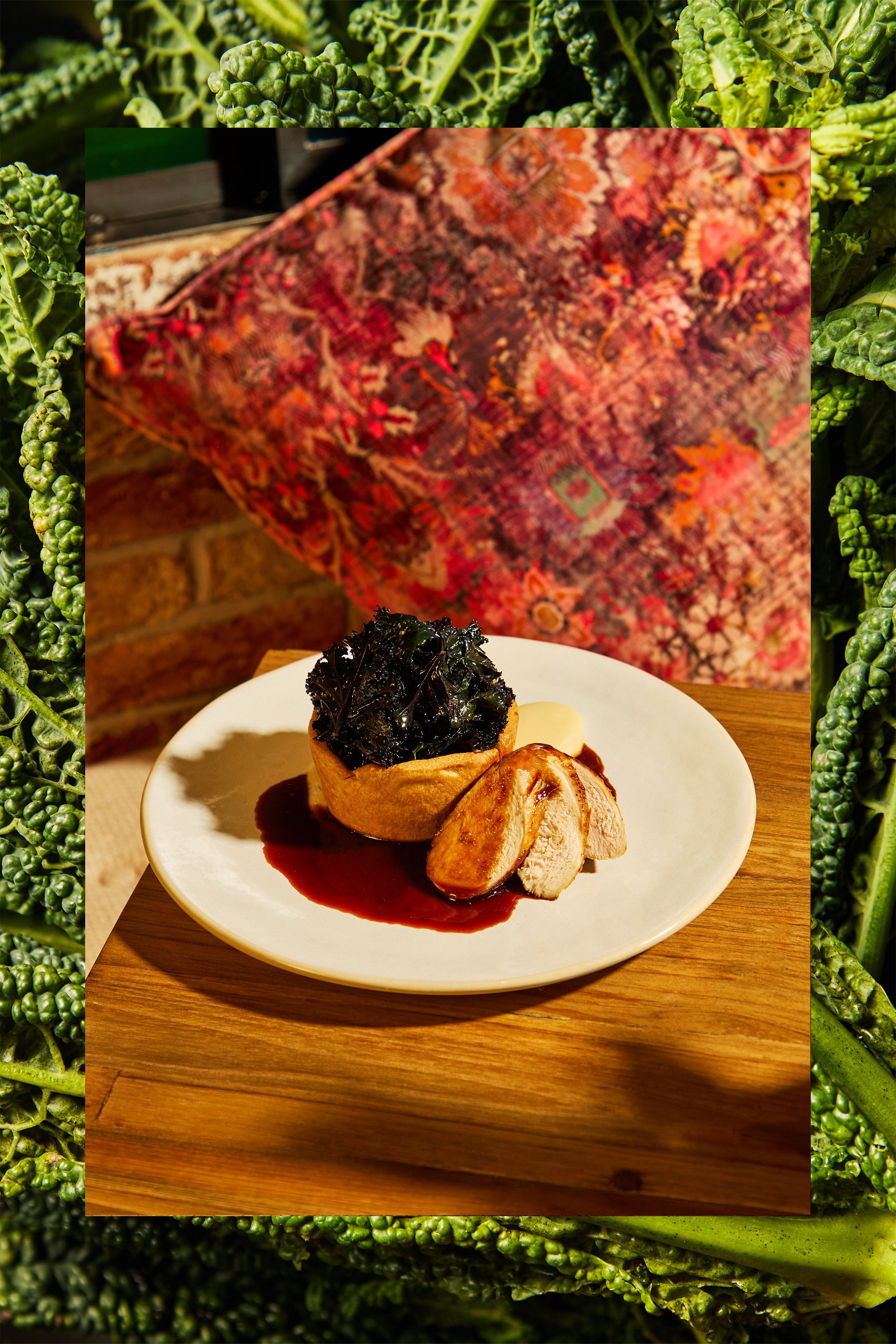 A plated gourmet meal with a pastry filled with dark leafy greens, sliced chicken, mashed potatoes, and a dark sauce, set on a wooden table with a colorful tapestry background and leafy green decor surrounding the image.