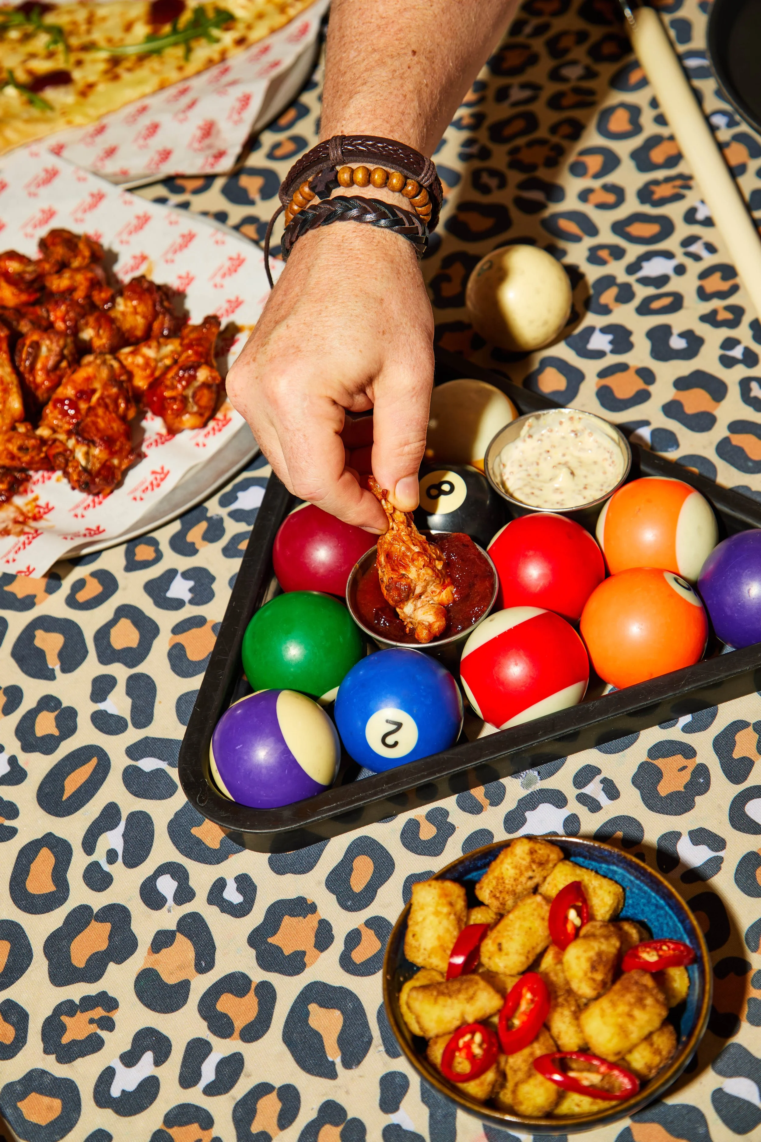 A person reaching for chicken wings with a pool ball in a black tray, surrounded by billiard balls, spicy chicken wings on a basket, and onion rings with red chili slices on a blue bowl, all on a leopard print tablecloth.