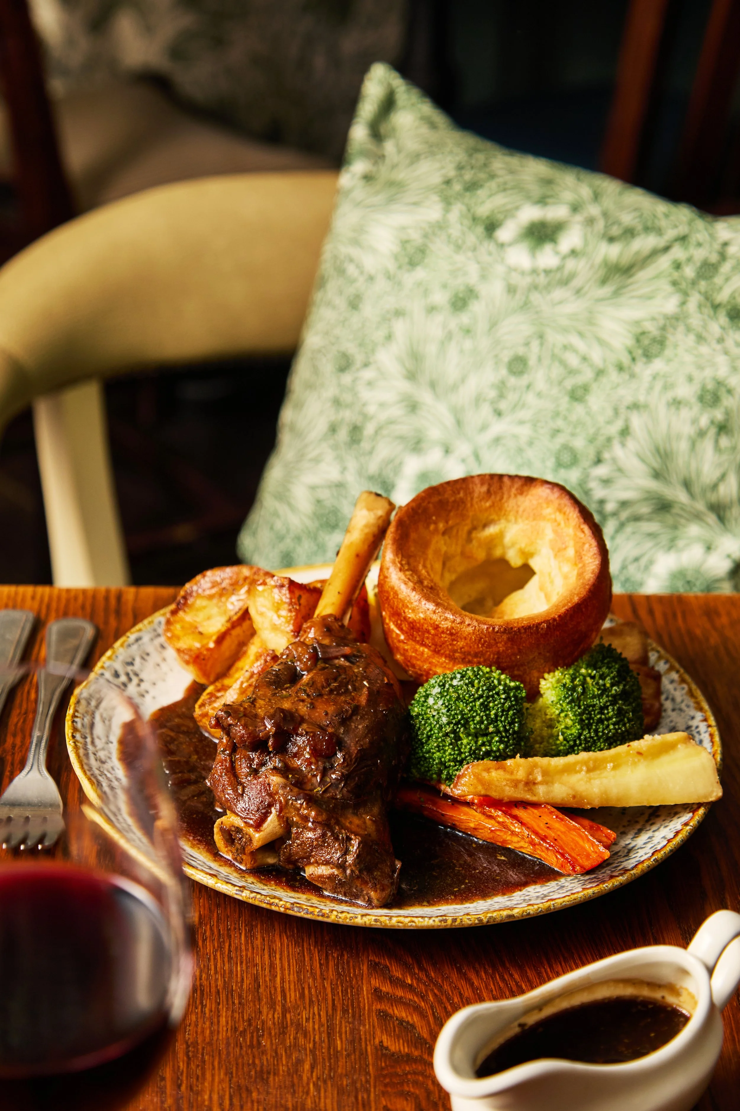 A plated meal featuring a large piece of meat with gravy, roasted potatoes, steamed broccoli, carrots, a slice of parsnip, and a bread bowl on a wooden table, with a glass of red wine and a small gravy boat.