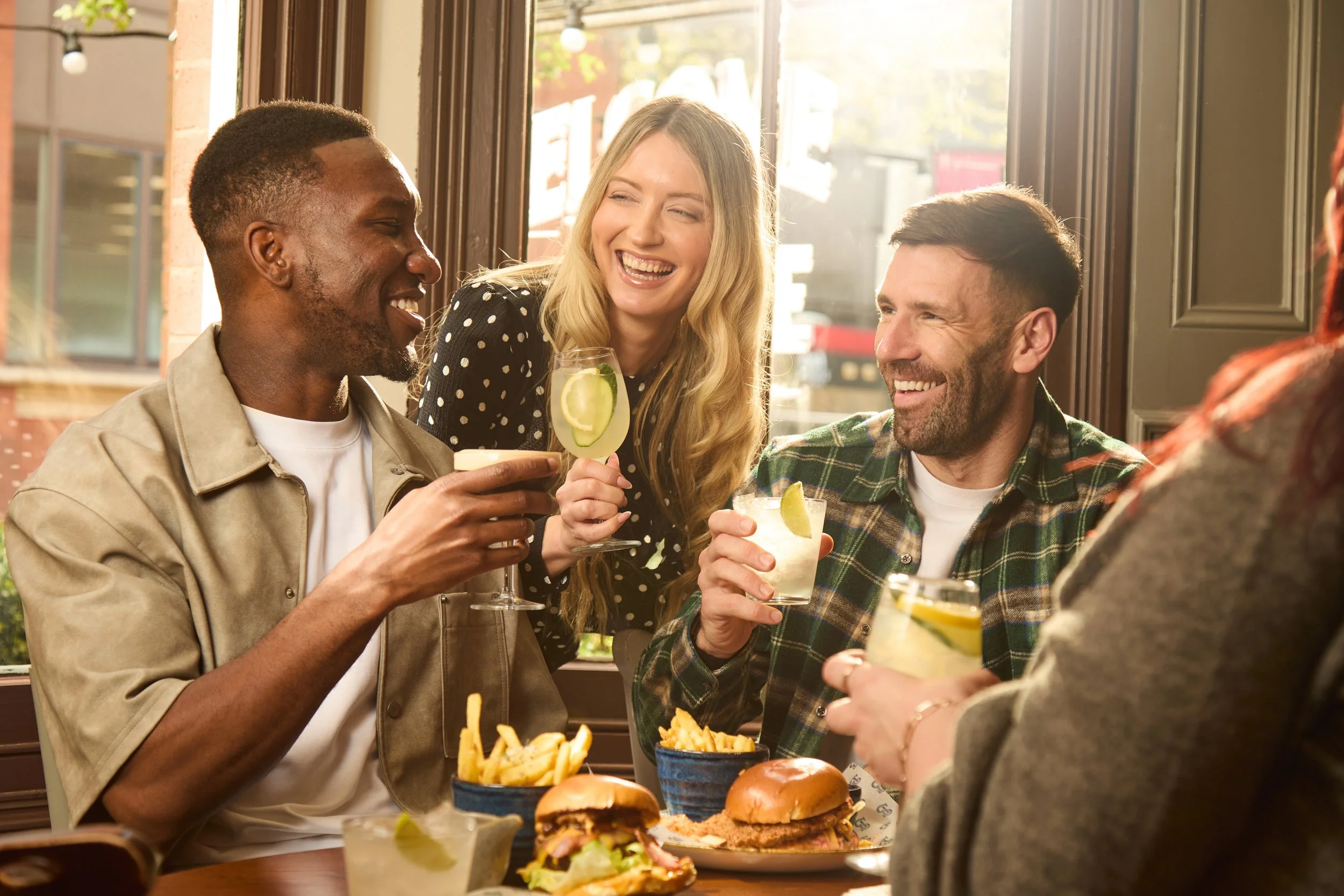 Group of four friends enjoying drinks and food at a restaurant, smiling and engaging with each other, with burgers, fries, and cocktails on the table.
