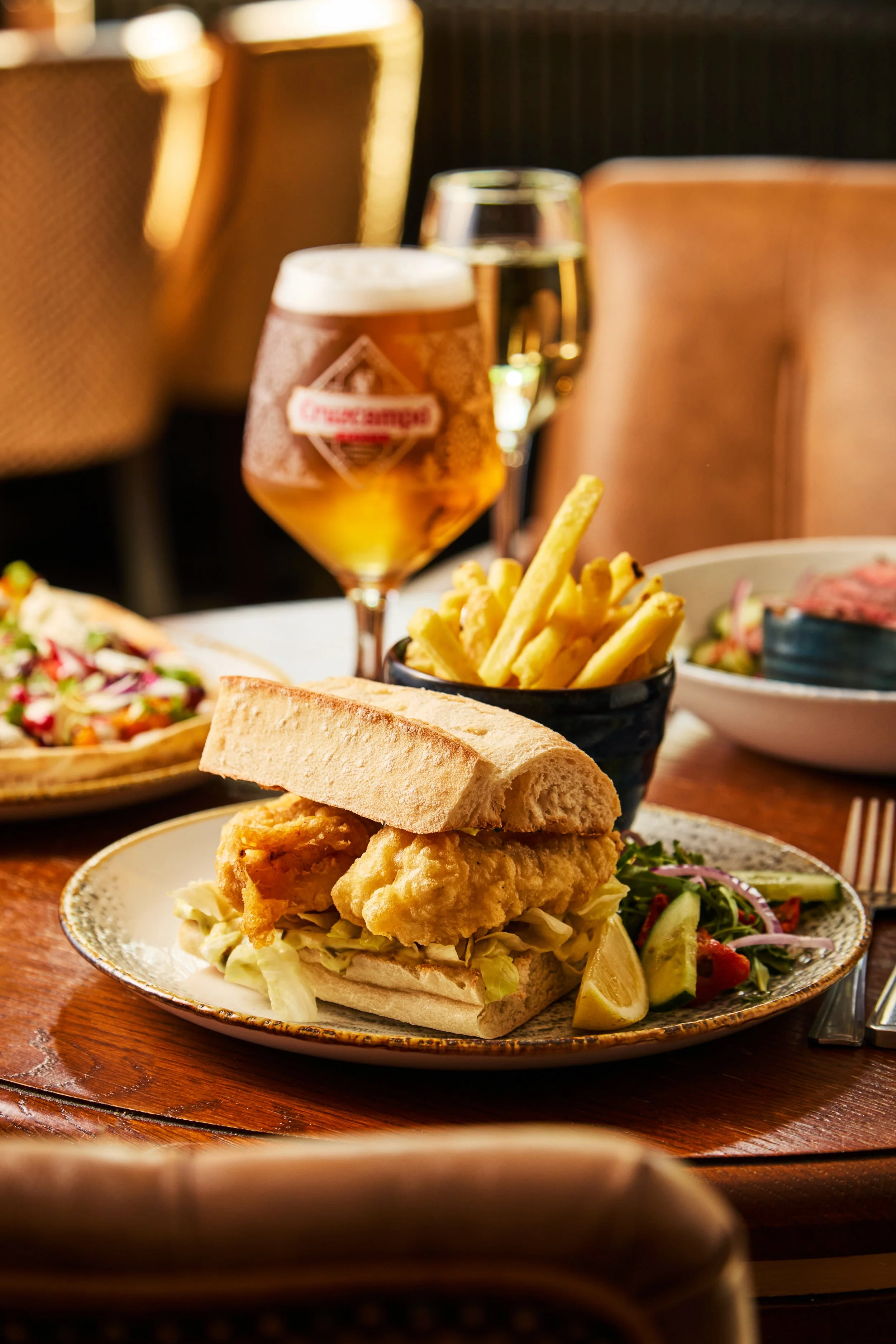 A plate with fried fish sandwich, shredded lettuce, and pickles, accompanied by a side salad. In the background, a glass of white wine and a glass of beer are visible on a wooden table.