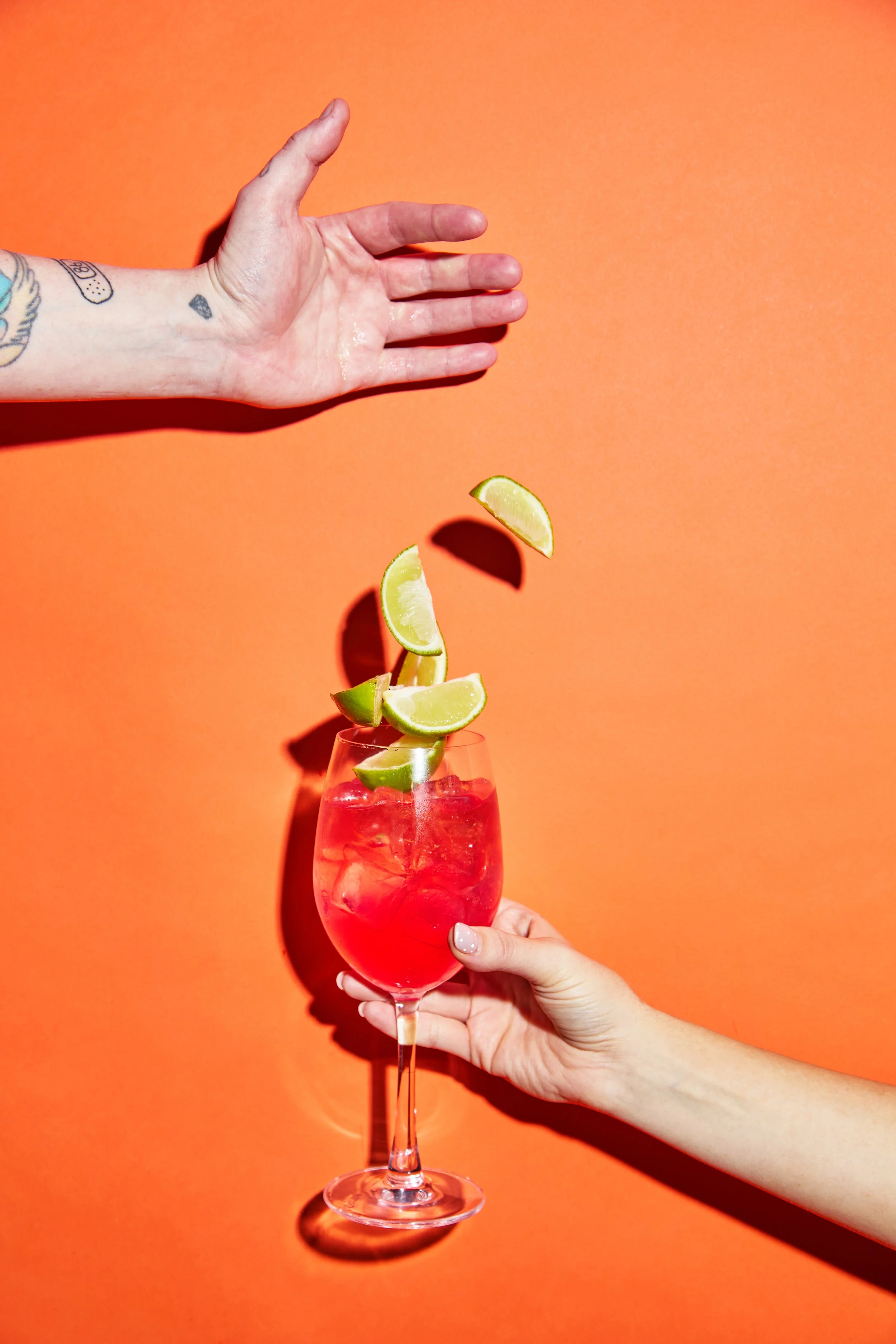 A person's hand is reaching towards a cocktail glass filled with red drink and ice, garnished with lime wedges falling into or out of the glass, against an orange background.