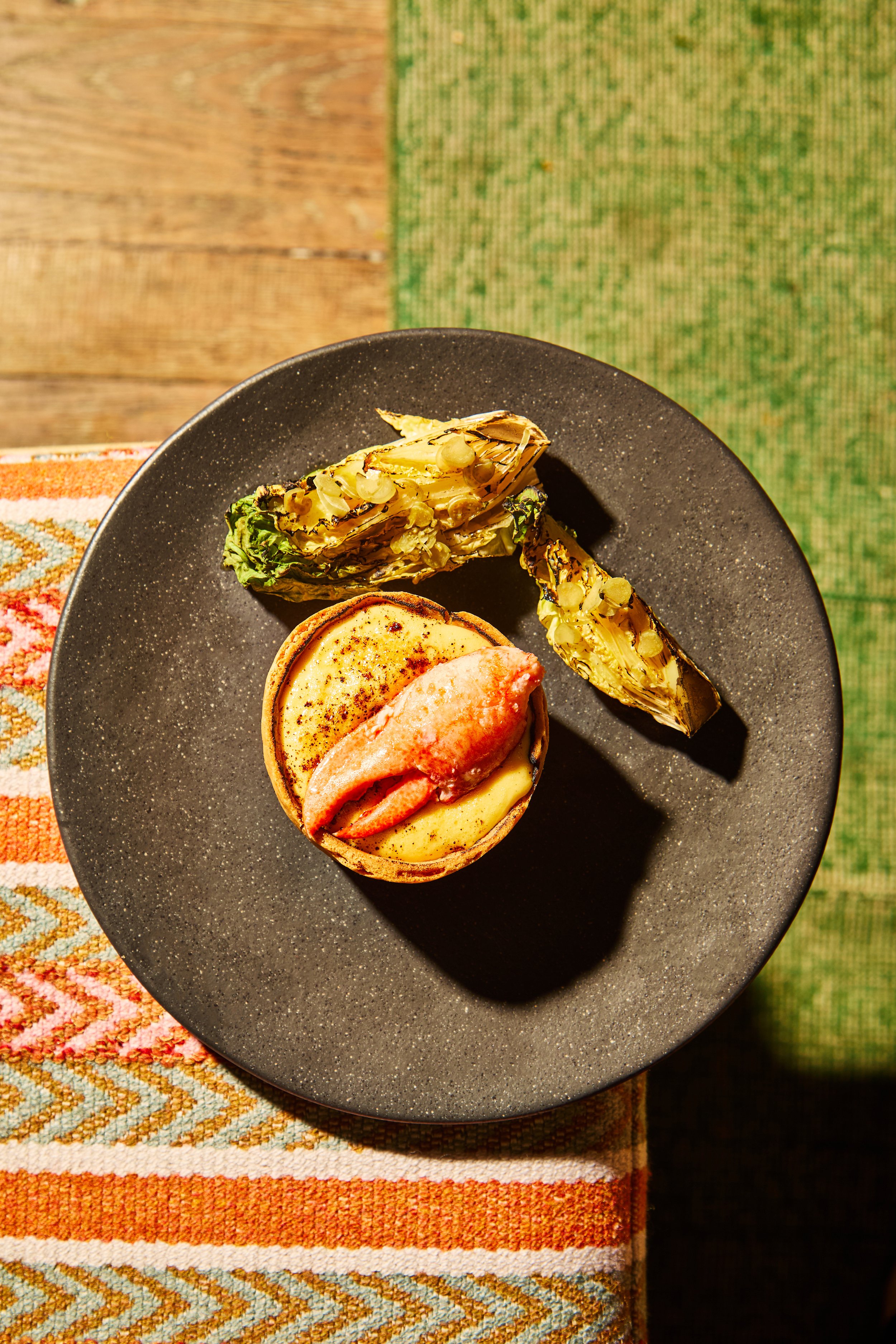 A dish of baked cheese with a slice of lobster, accompanied by roasted artichoke halves, served on a dark plate placed on a colorful tablecloth.