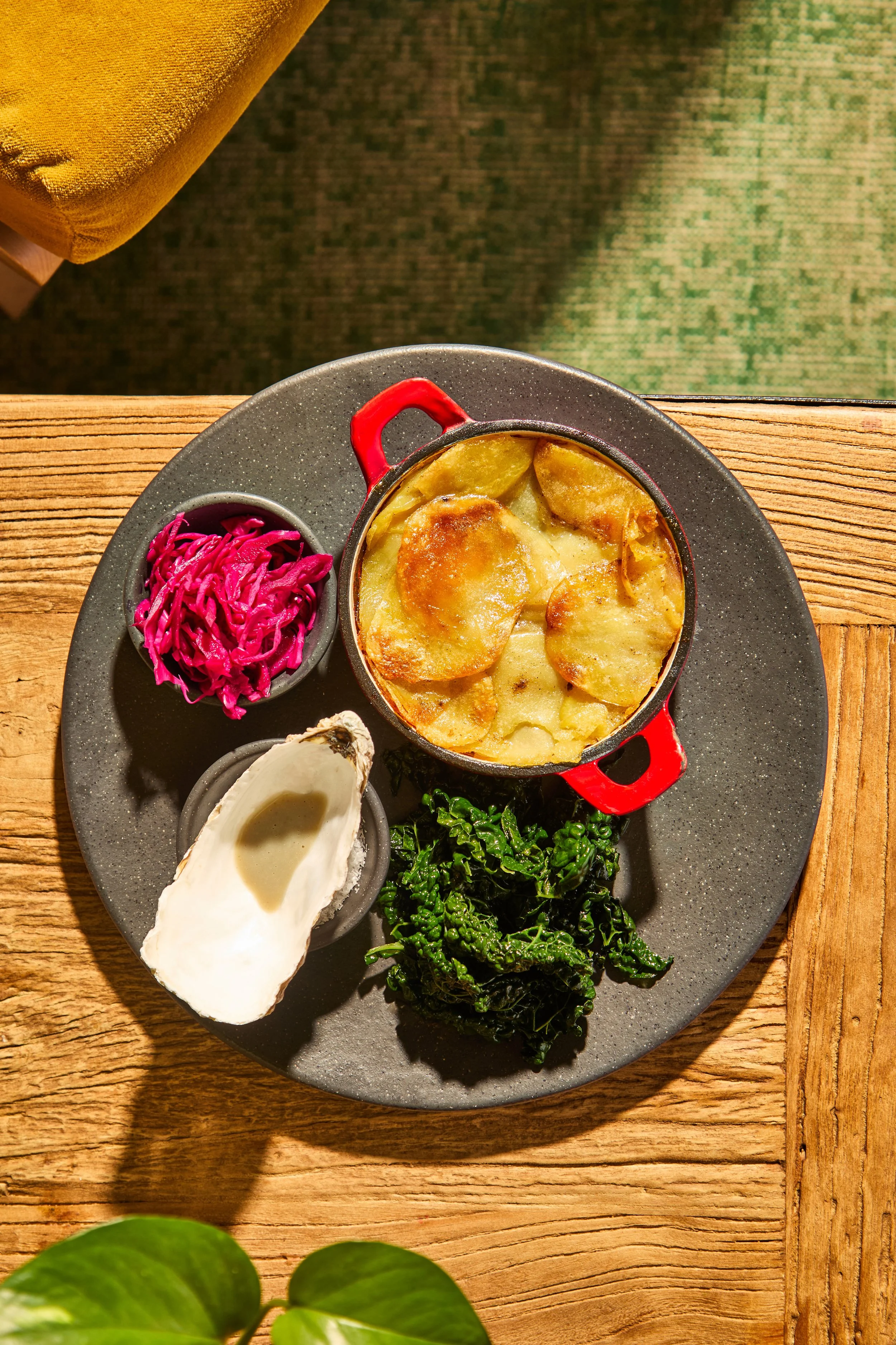 A round plate with a small cast iron skillet of scalloped potatoes, a bowl of pink pickled cabbage, a bowl of raw oysters, and a portion of sautéed kale, on a wooden table.