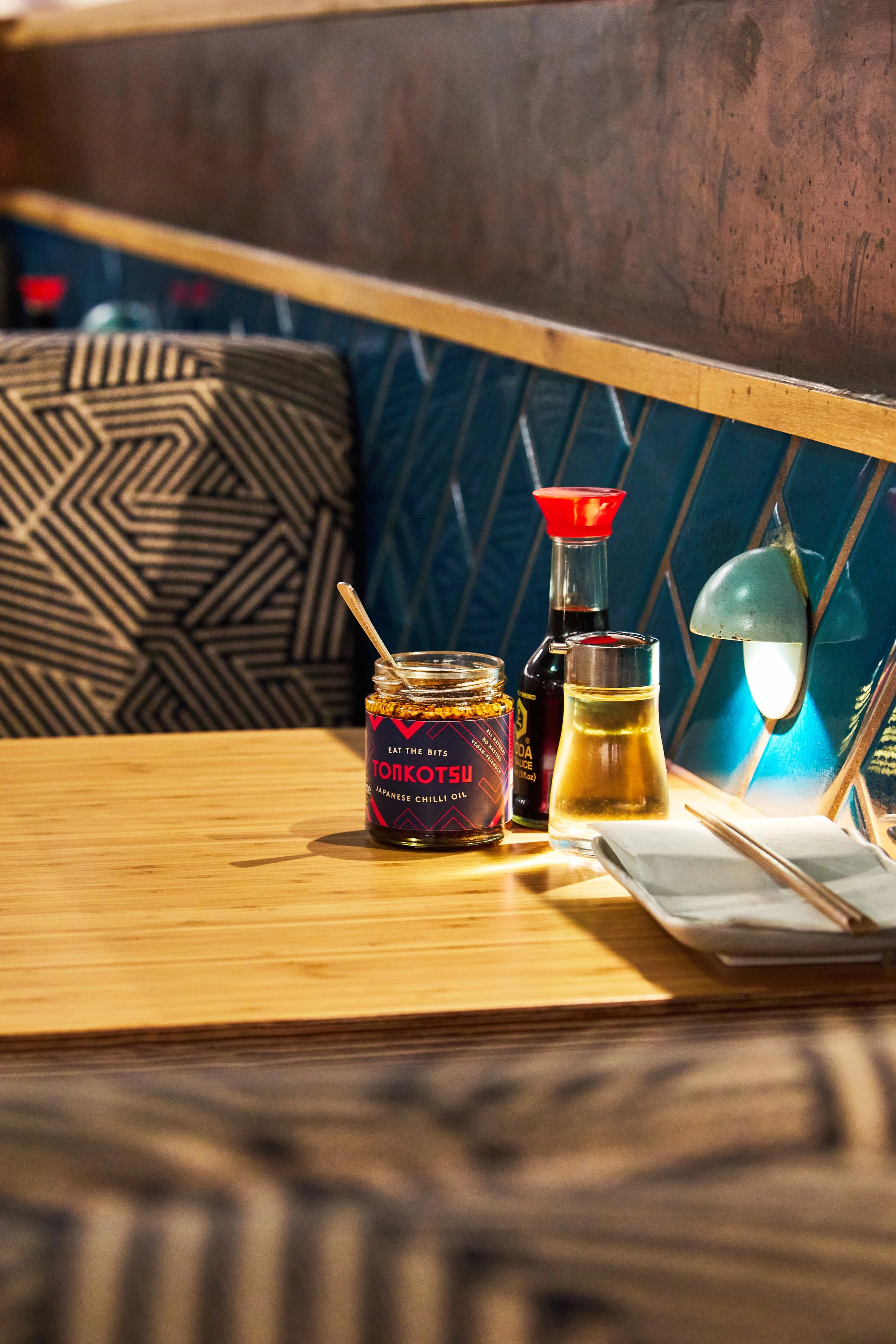 A restaurant table with a jar of Japanese chili oil, a soy sauce bottle, a small tray with chopsticks, and condiments, against a blue tiled wall and a cushioned seat in the background.