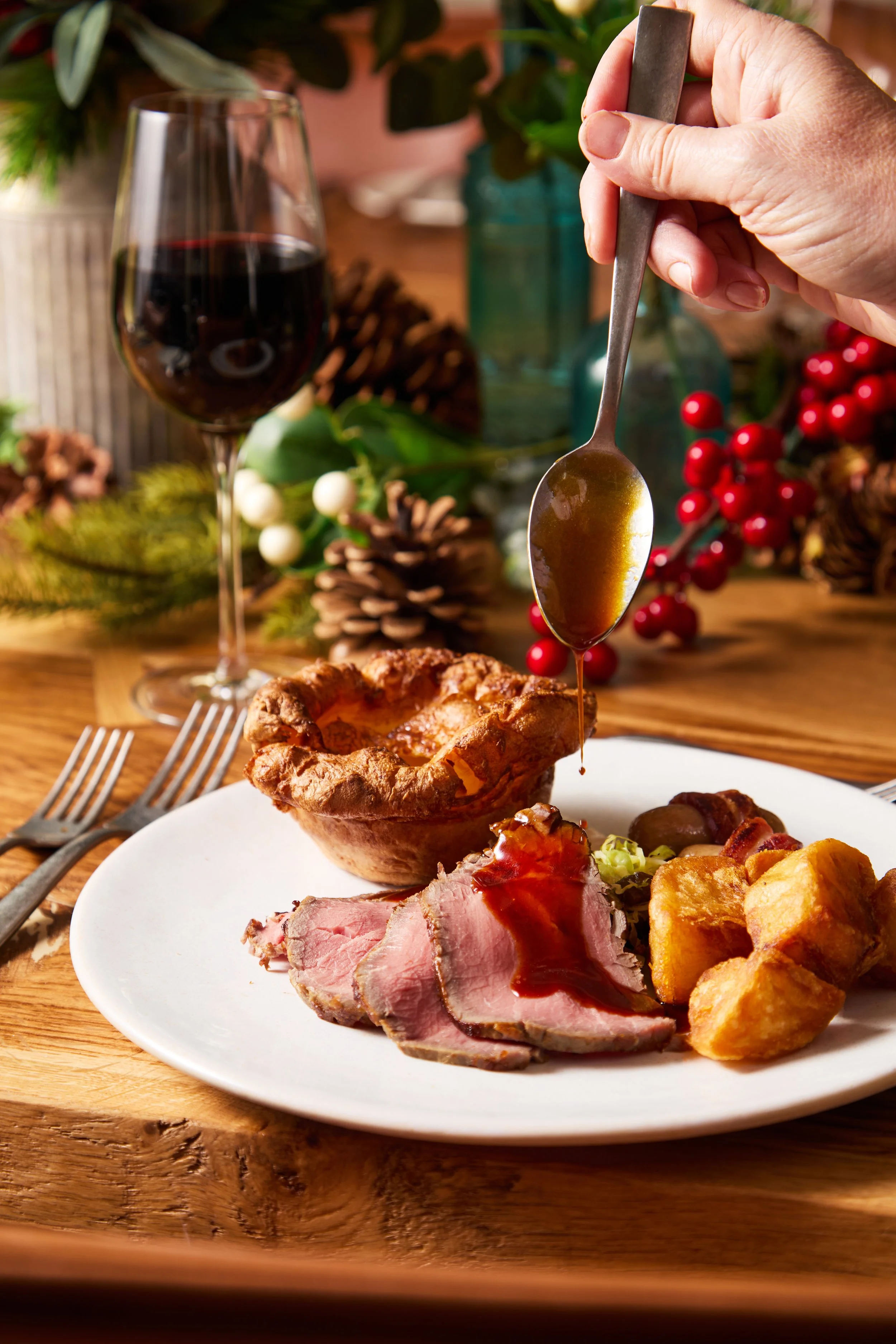 A Christmas dinner plate with sliced ham, Yorkshire pudding, roasted potatoes, and vegetables, with a glass of red wine and festive decorations in the background.