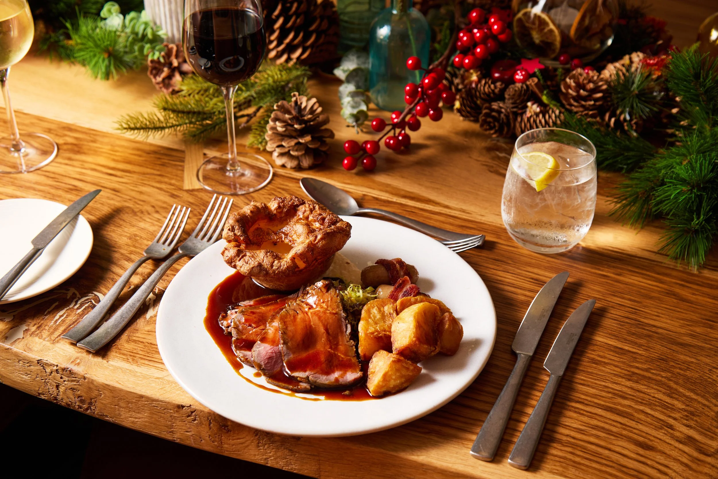 A holiday dinner table with a plate of roast meat, potatoes, Yorkshire pudding, and vegetables, surrounded by holiday decorations including pine branches, pinecones, red berries, and greenery, with glasses of red wine and water.