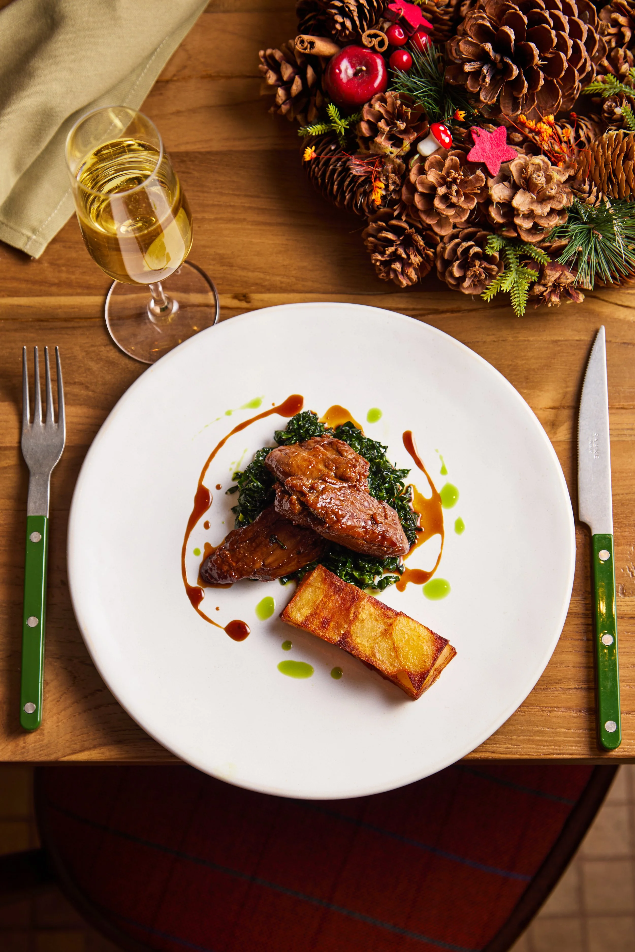 A plated dish featuring braised meat, sautéed greens, crispy potato stack, garnished with sauce and oil drops, on a white plate beside a glass of white wine, on a wooden table decorated with a Christmas centerpiece