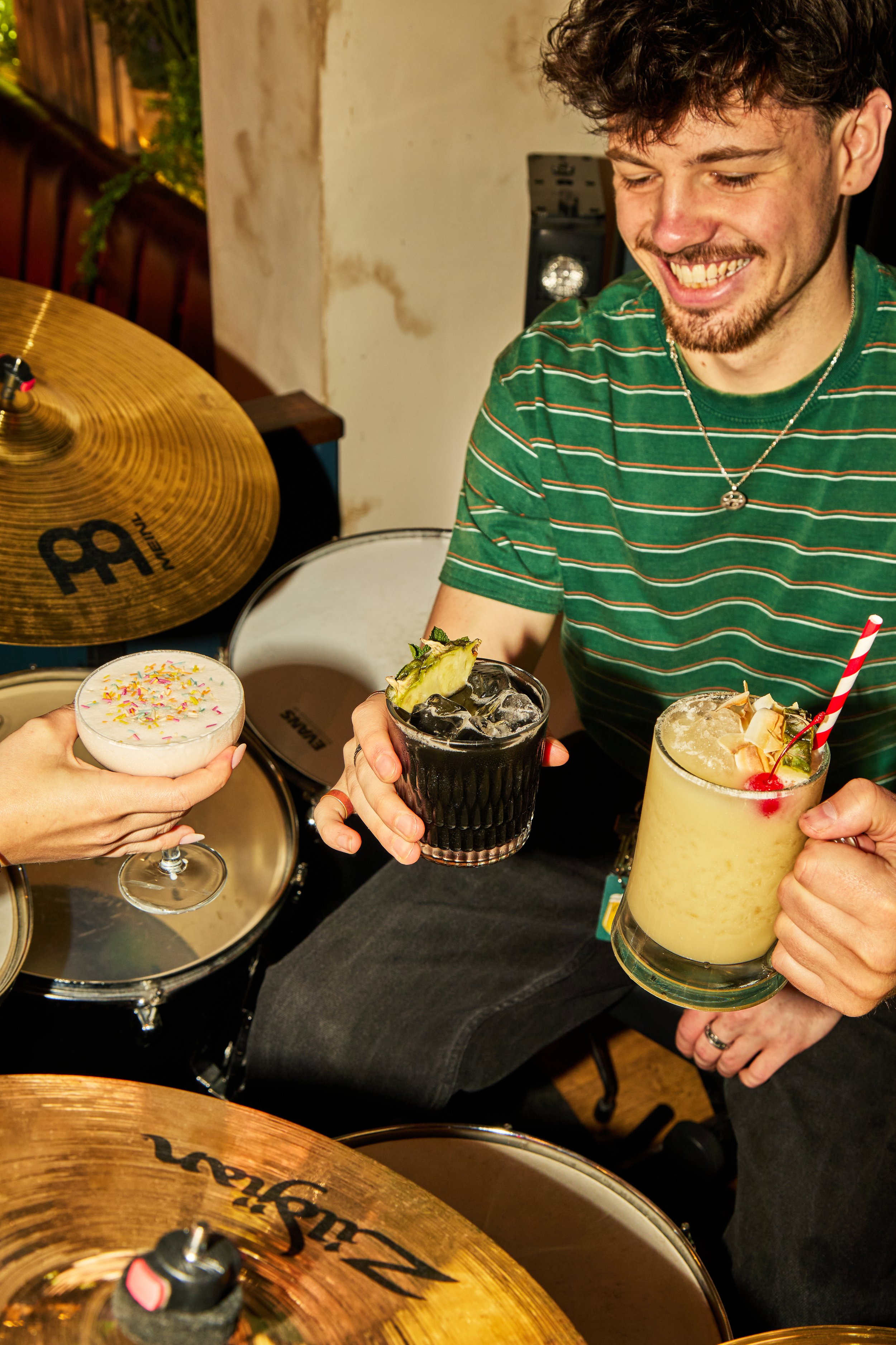 A person with curly hair, wearing a green striped shirt and a necklace, smiling and holding three different cocktails: one with whipped cream and sprinkles, a dark drink with pineapple and mint, and a yellow drink with a cherry, pineapple, and coconu