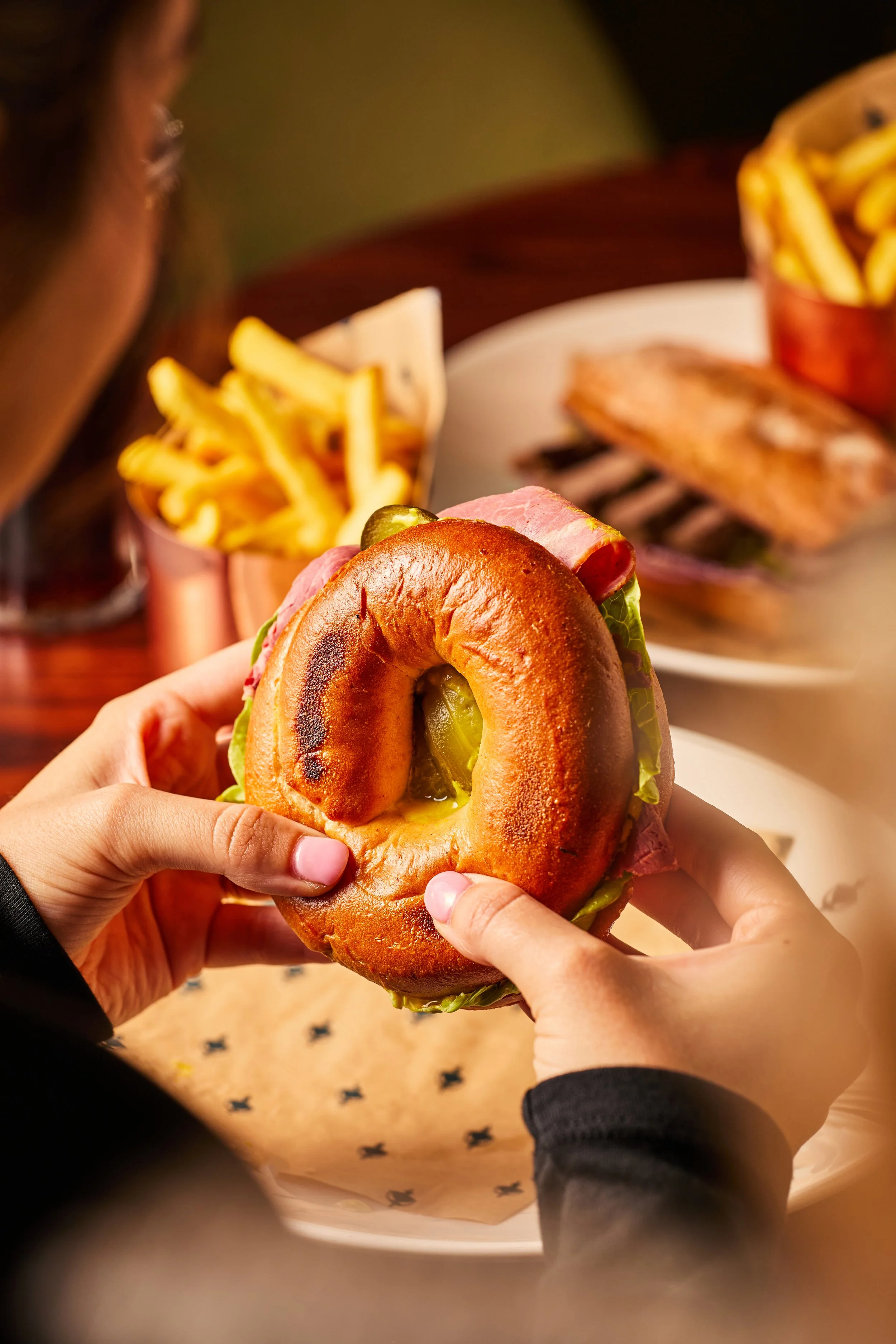 Person holding a bagel sandwich with lettuce, pickles, ham, and mustard. French fries in copper cups and grilled fish on a white plate in the background.