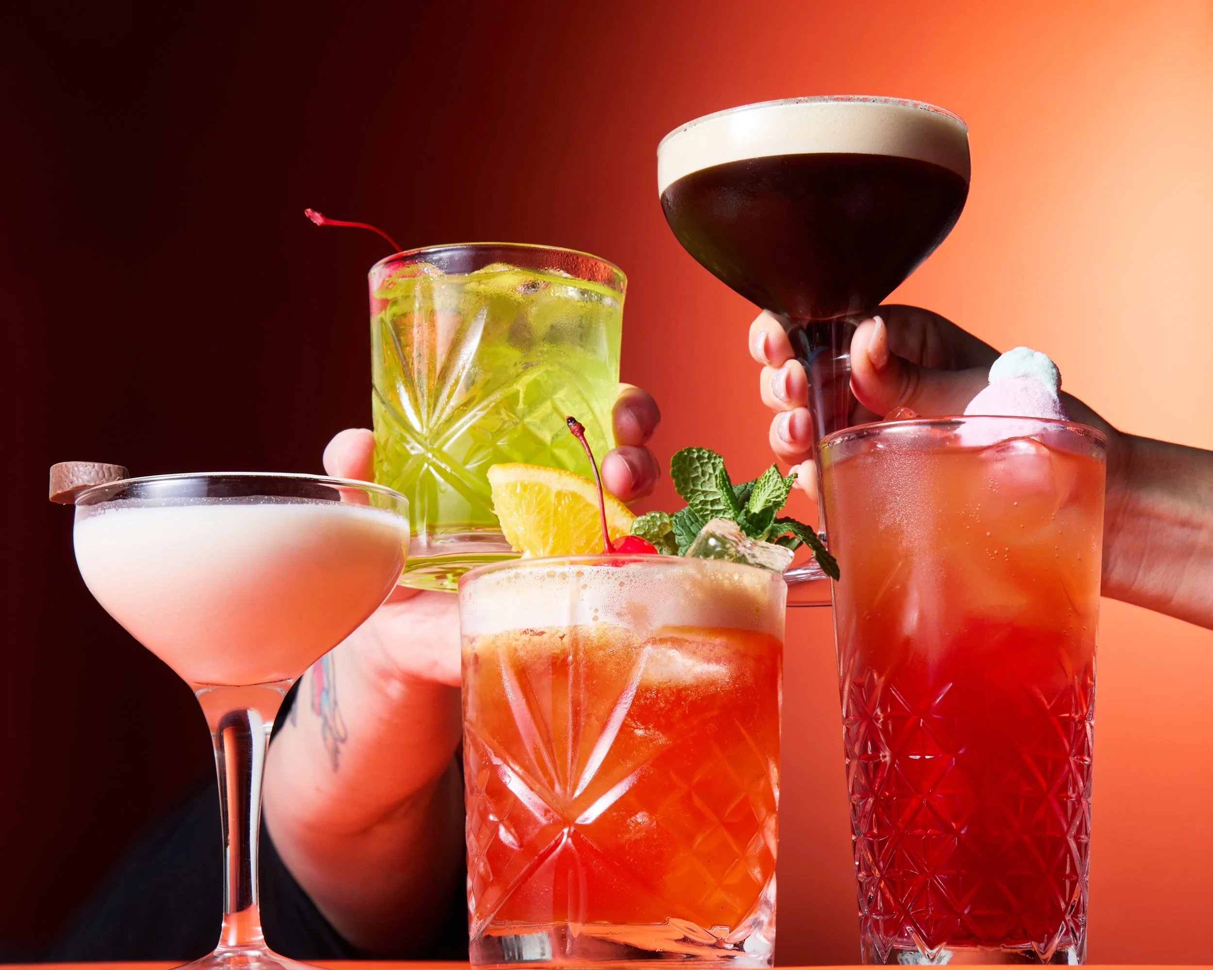 A variety of colorful cocktails and drinks with garnishes such as cherries, lemon slices, and mint leaves, held by two hands against a red and orange background.