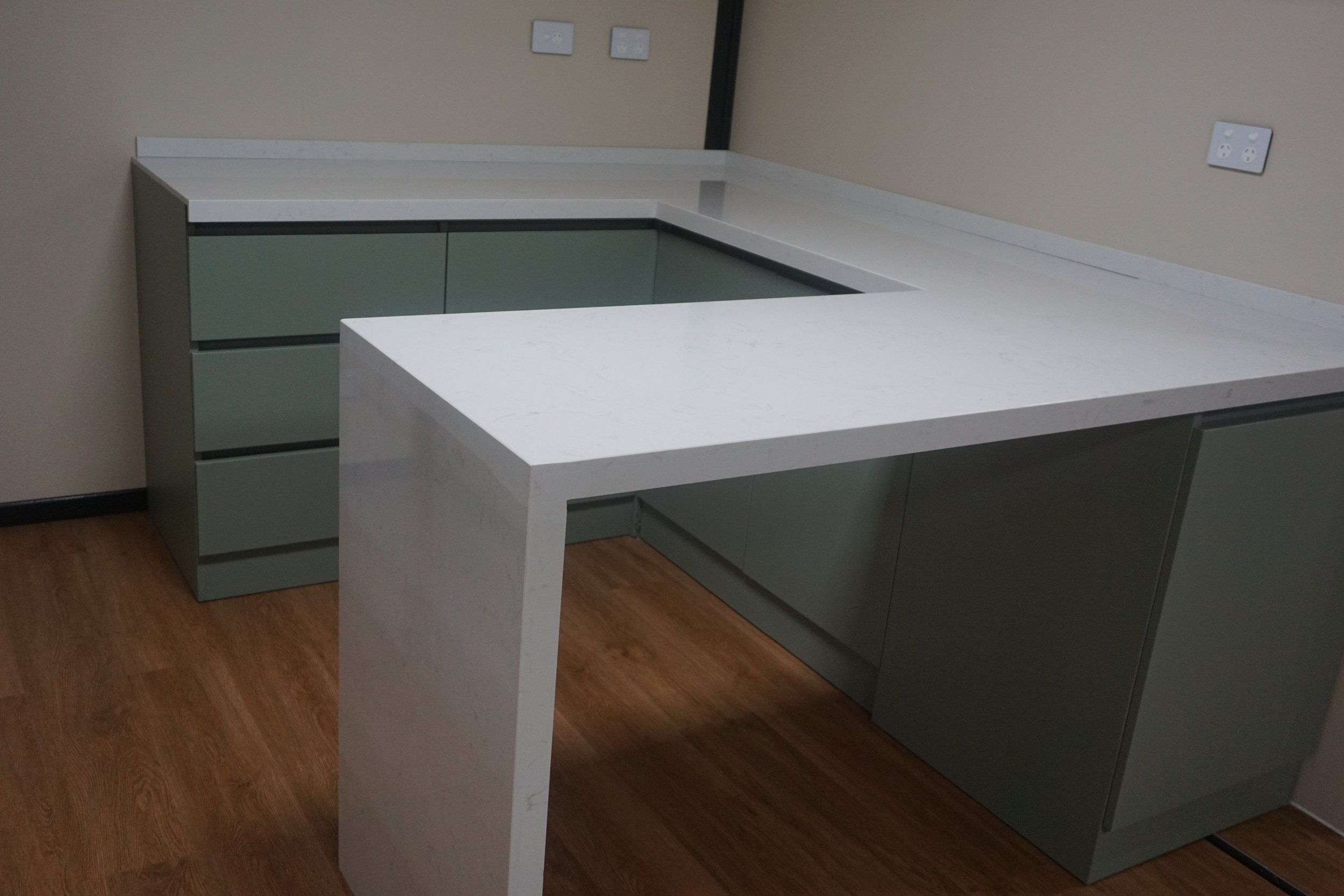 Empty L-shaped office desk with a white marble top and green cabinets against a beige wall with electrical outlets