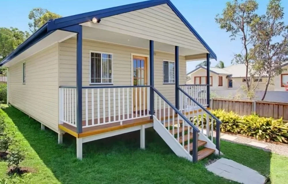 Front porch of a small, white, wooden house with blue trim, stairs leading up to the entrance, and a small yard with grass and plants.