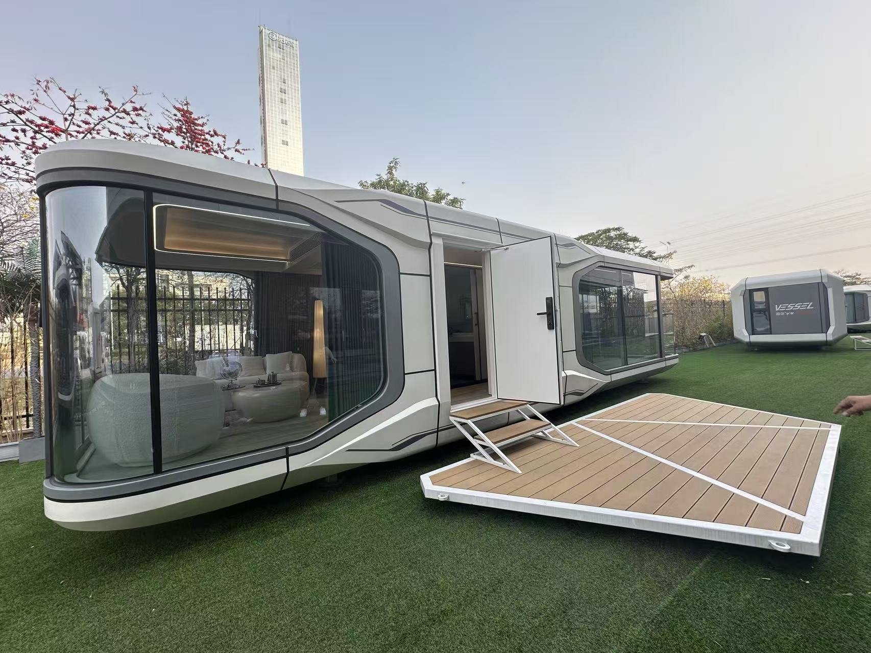 Modern futuristic tiny house on wheels with large glass windows, an open door with stairs, and a wooden deck, set outdoors on grass with a small additional building in the background.