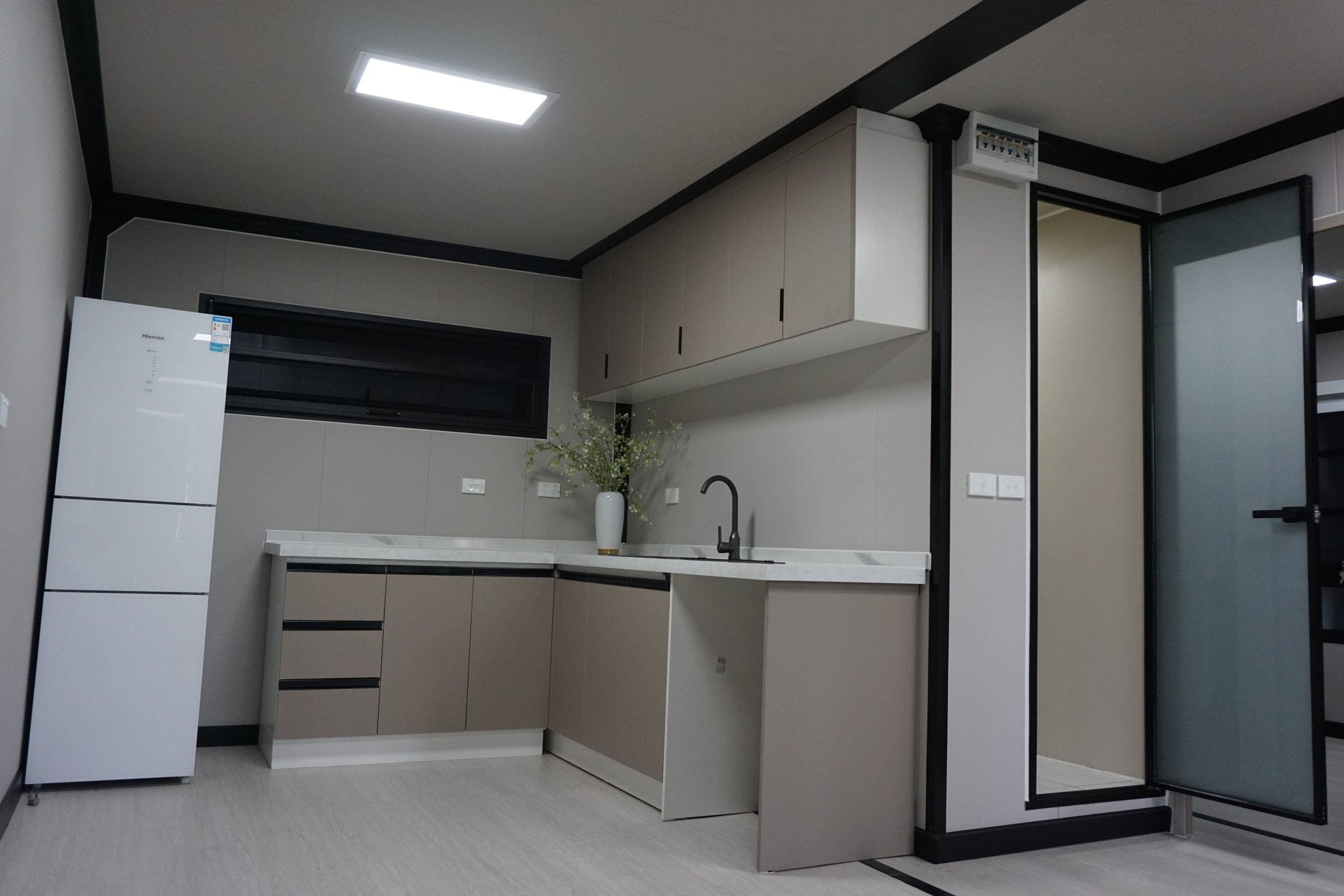 Modern kitchen with beige cabinets, marble countertop, black faucet, white vase with flowers, stainless steel refrigerator, window, and glass door.