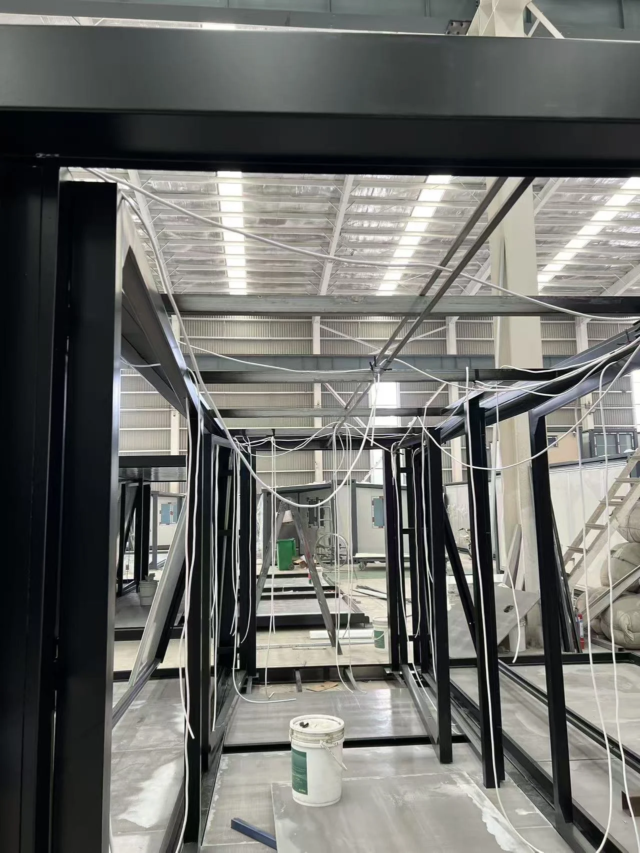 Industrial construction site with black metal frame structures, white electrical wires hanging loosely, and a white bucket in the foreground, inside a warehouse or factory with a high ceiling and metal roof.