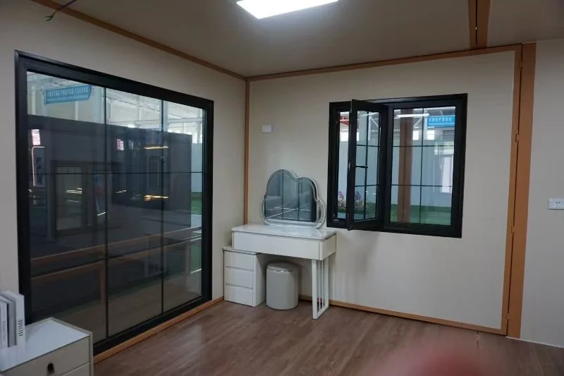 A room with hardwood floors, a vanity with a mirror, a small white drawer, and a trash bin underneath the vanity. There are two black-framed windows, one sliding door, and a ceiling light.