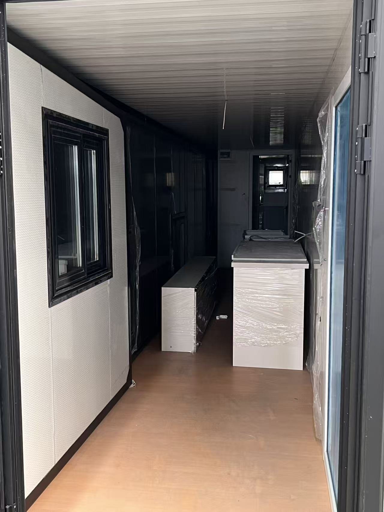 Interior of a mobile home or trailer with black cabinets, white countertops, and a wood laminate floor, with plastic wrapping on some surfaces.