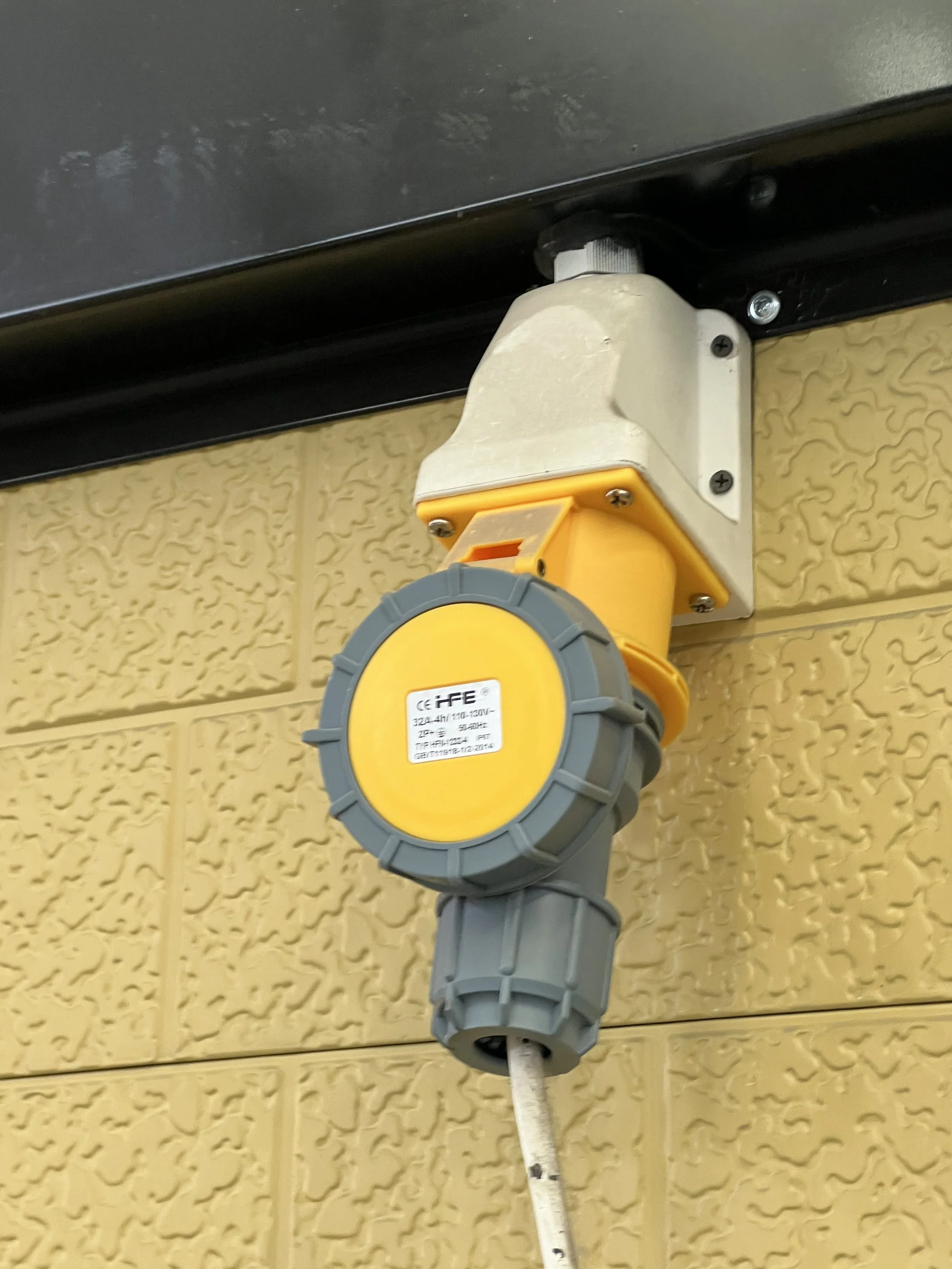 Industrial electrical outlet with a gray and yellow cover attached to a beige brick wall