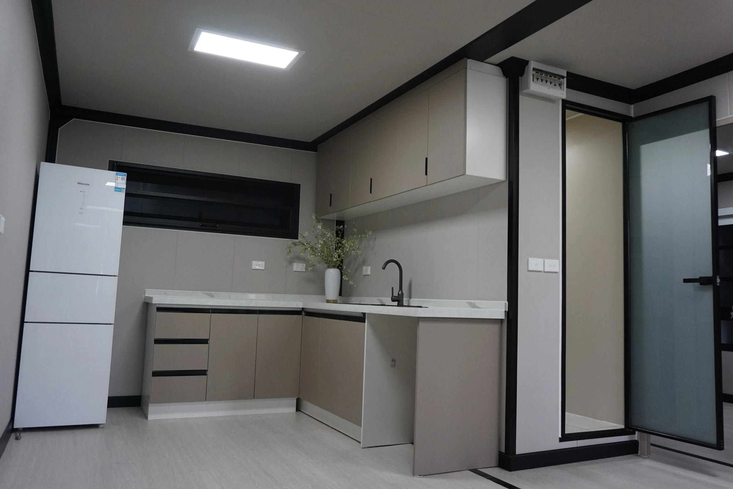 Modern kitchen with beige cabinets, white marble countertop, black faucet, white vase with flowers, refrigerator, and a window.