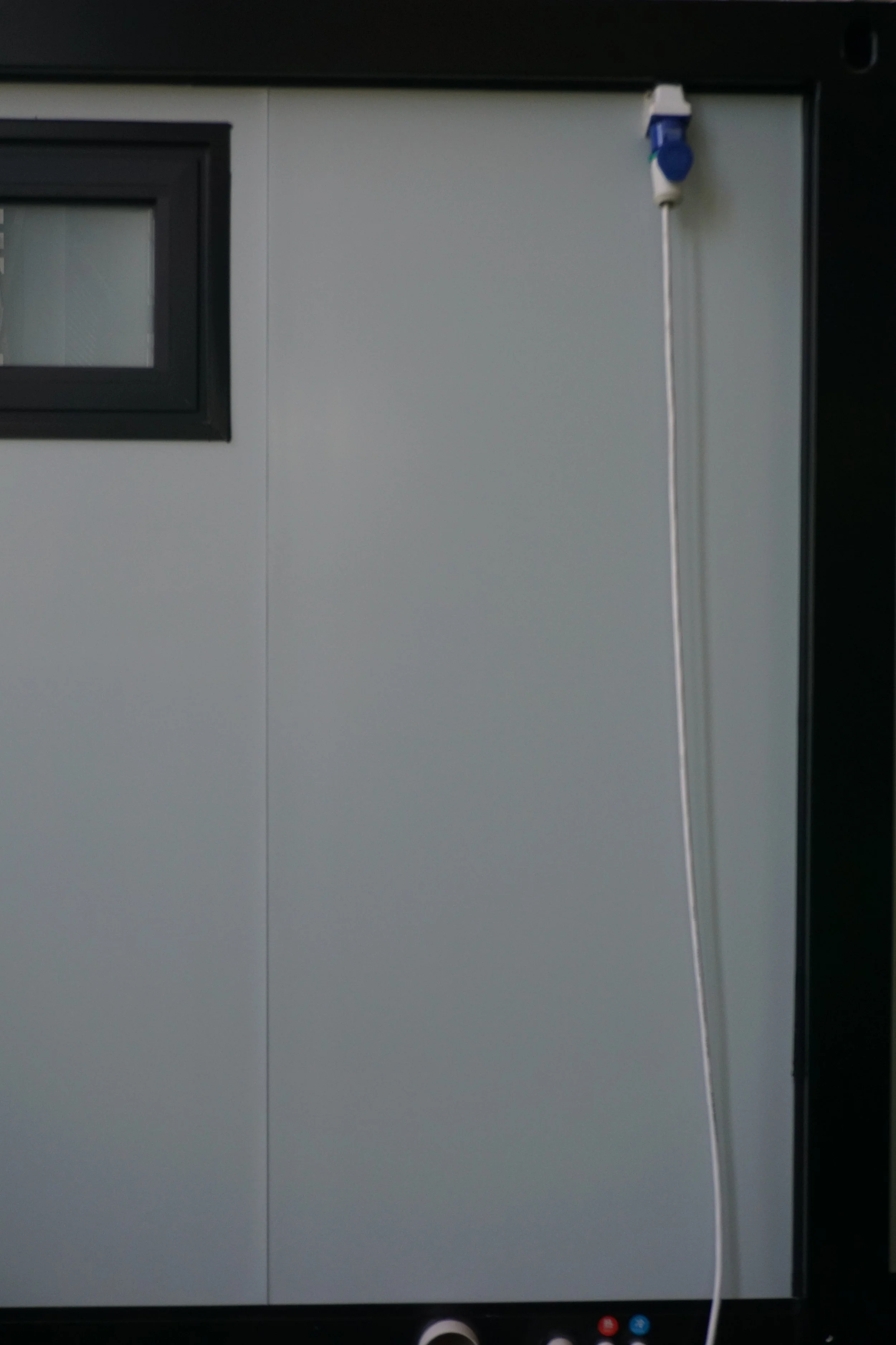 Close-up of a gray panel with a small, black-framed window in the upper left corner and a white cable hanging beside a blue and white socket in the upper right corner.