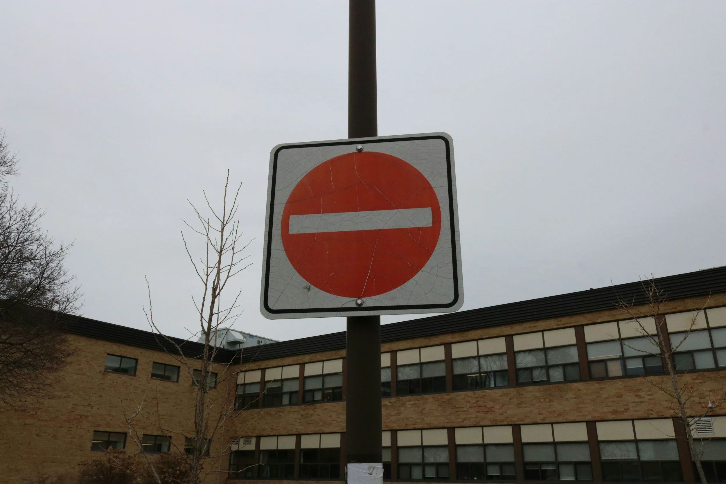 A red “No Entry” road sign positioned outside a school building, symbolising restricted access and questioning traditional education pathways for neurodivergent children.