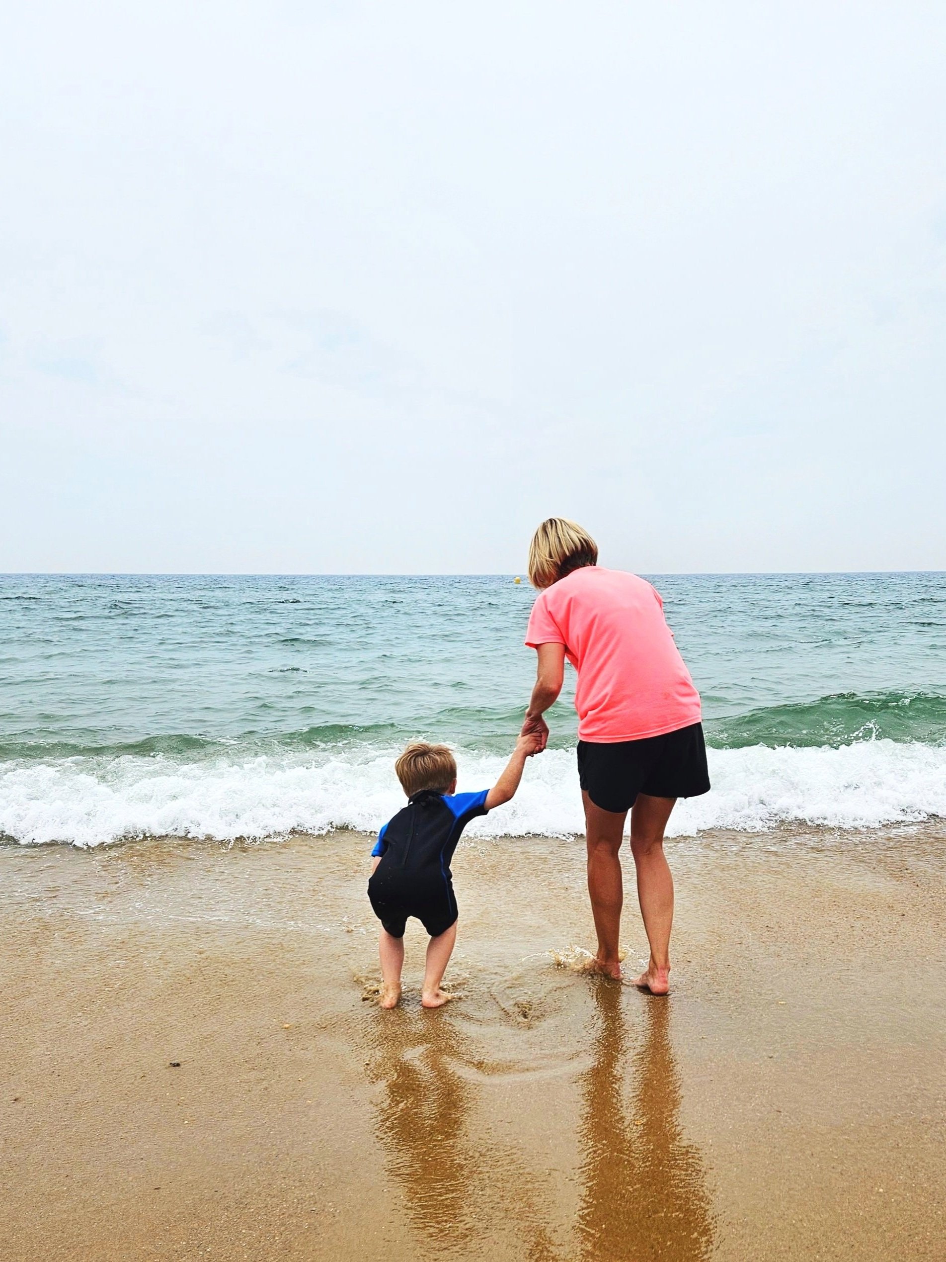 Mother and son standing on a beach with their backs to the camera, holding hands and looking out at the sea, symbolising support, protection and parental advocacy.