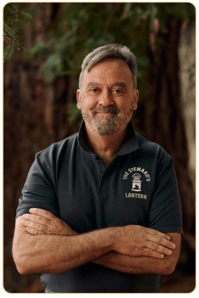 Image of Andrew Lockhart McLachlan standing under a pine tree. His arms are folded, smile on his face. He is wearing a charcoal grey polo shirt with The Steward's Lantern logo embroidered on the left side.